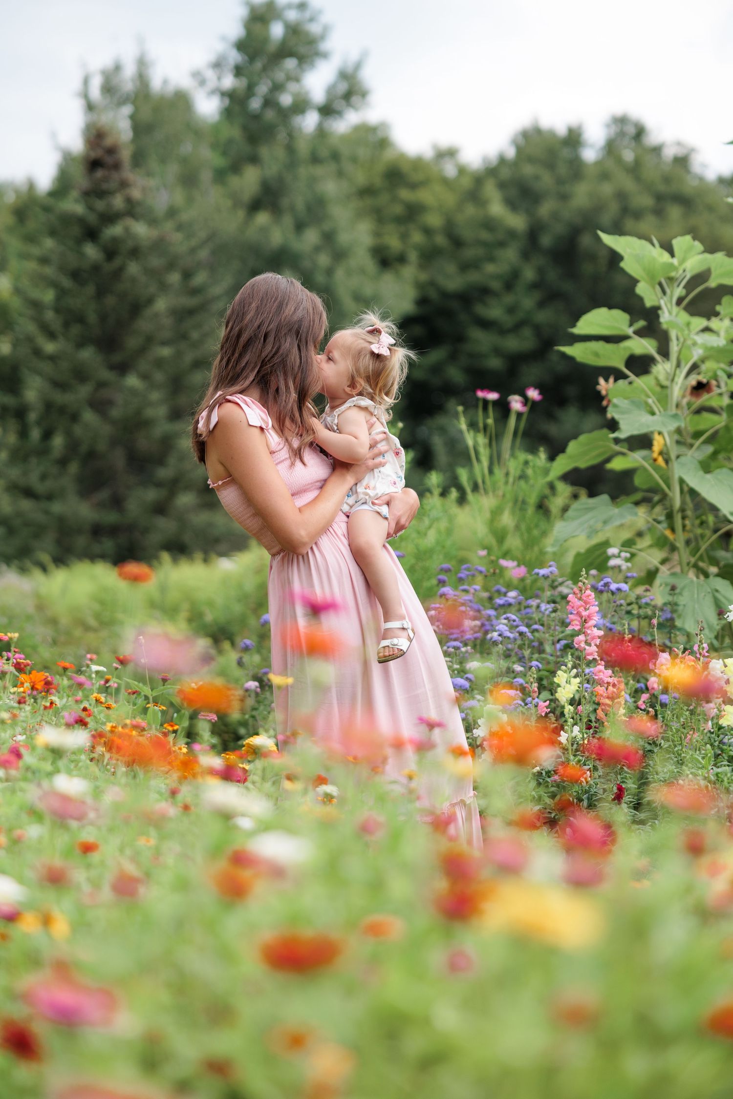 A mother kisses her young daughter while standing in a vibrant field of colorful flowers.