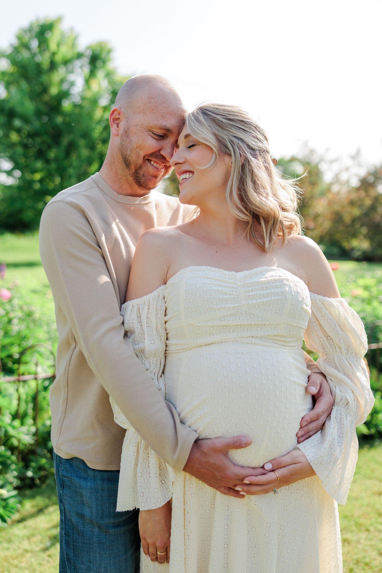 Loving couple cuddling outdoors, man embracing pregnant woman in white off-shoulder dress in green park.