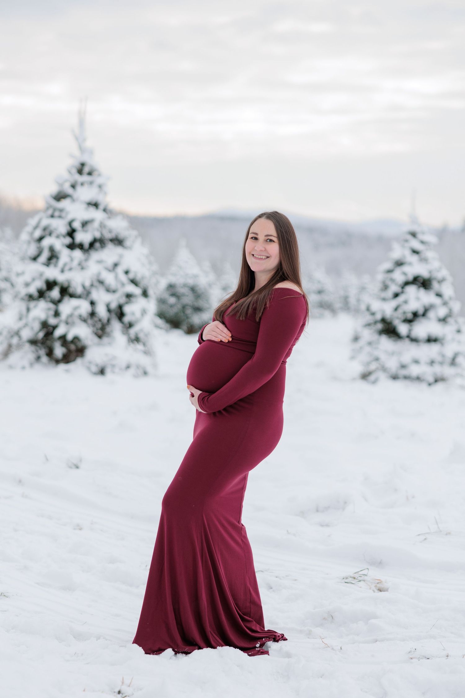 A pregnant woman in a burgundy gown stands confidently among snow-covered trees in a winter landscape.