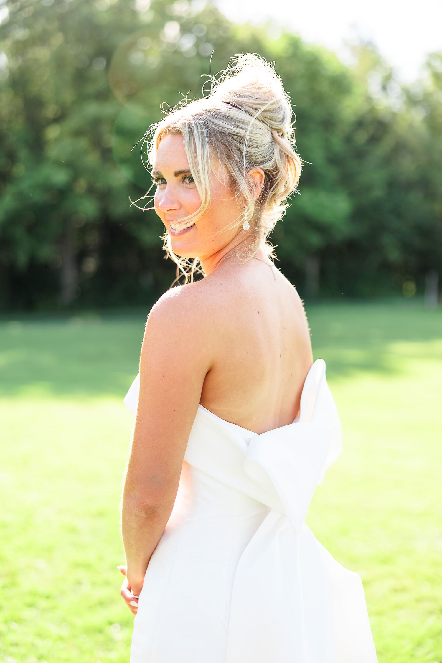 Bride in strapless white gown with bow back smiles over shoulder during outdoor bridal portrait session.