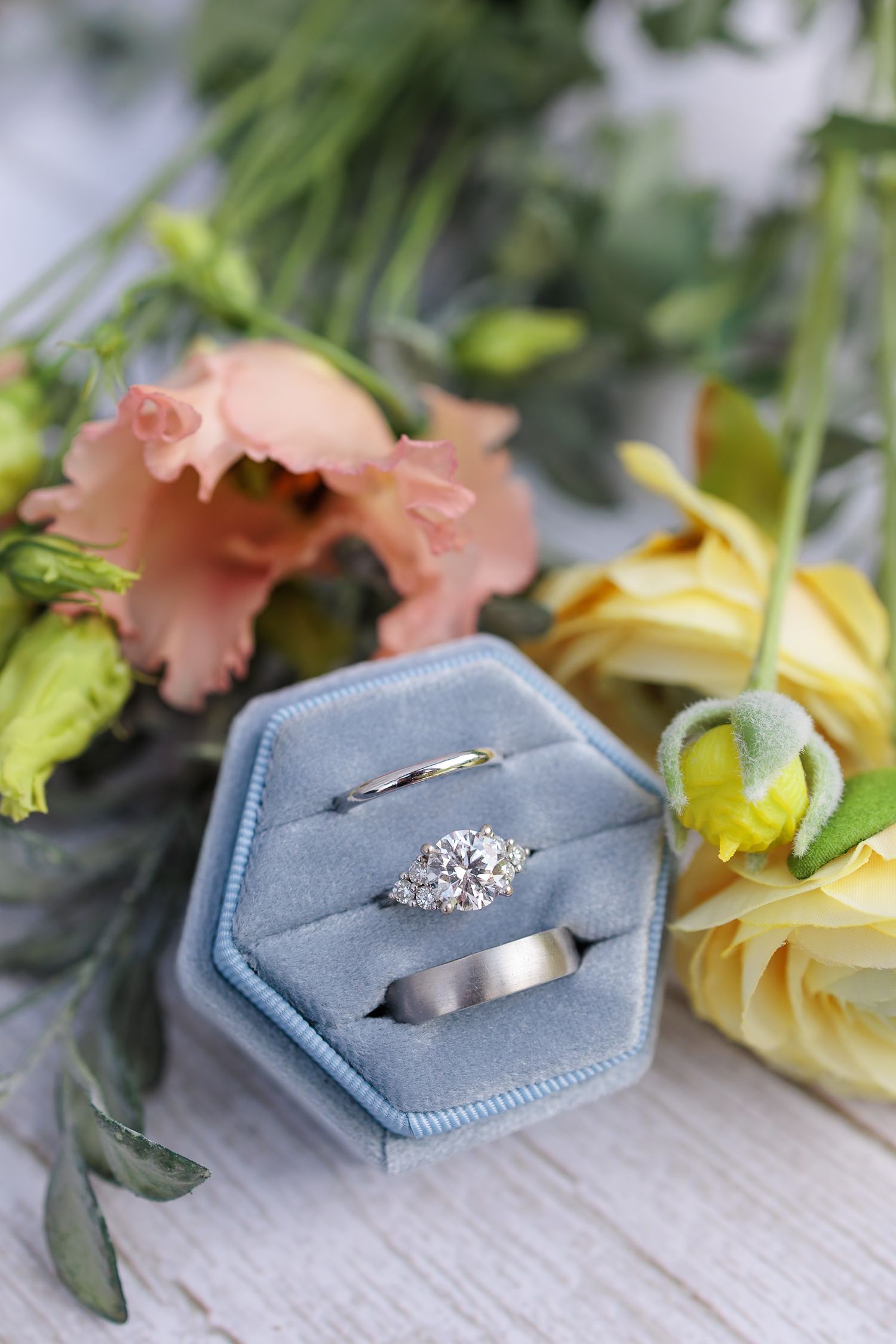 Diamond engagement ring and wedding bands displayed in blue velvet hexagon box with pink and yellow flowers.