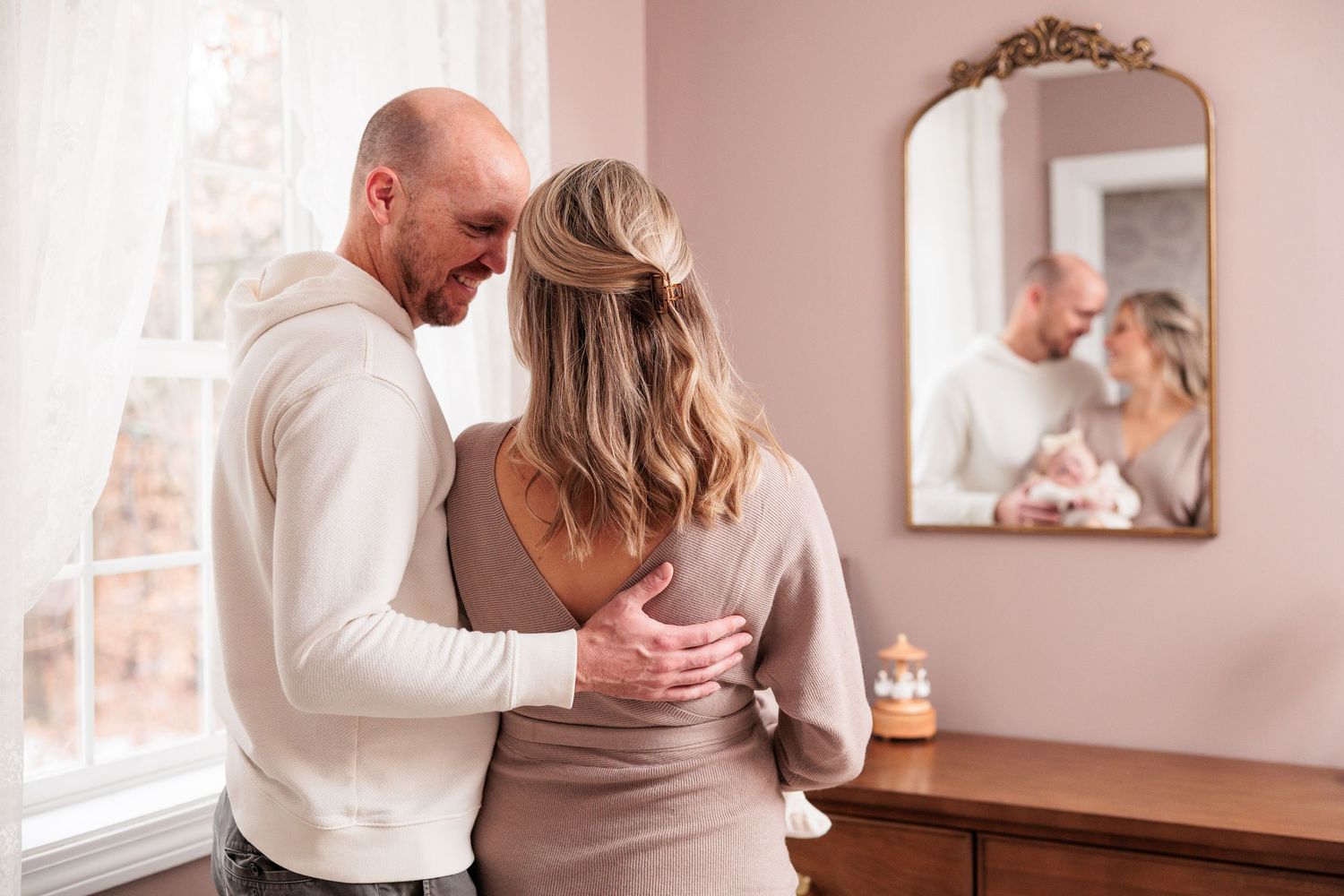 A couple dancing intimately in a softly lit room with pink walls and natural light streaming through windows.