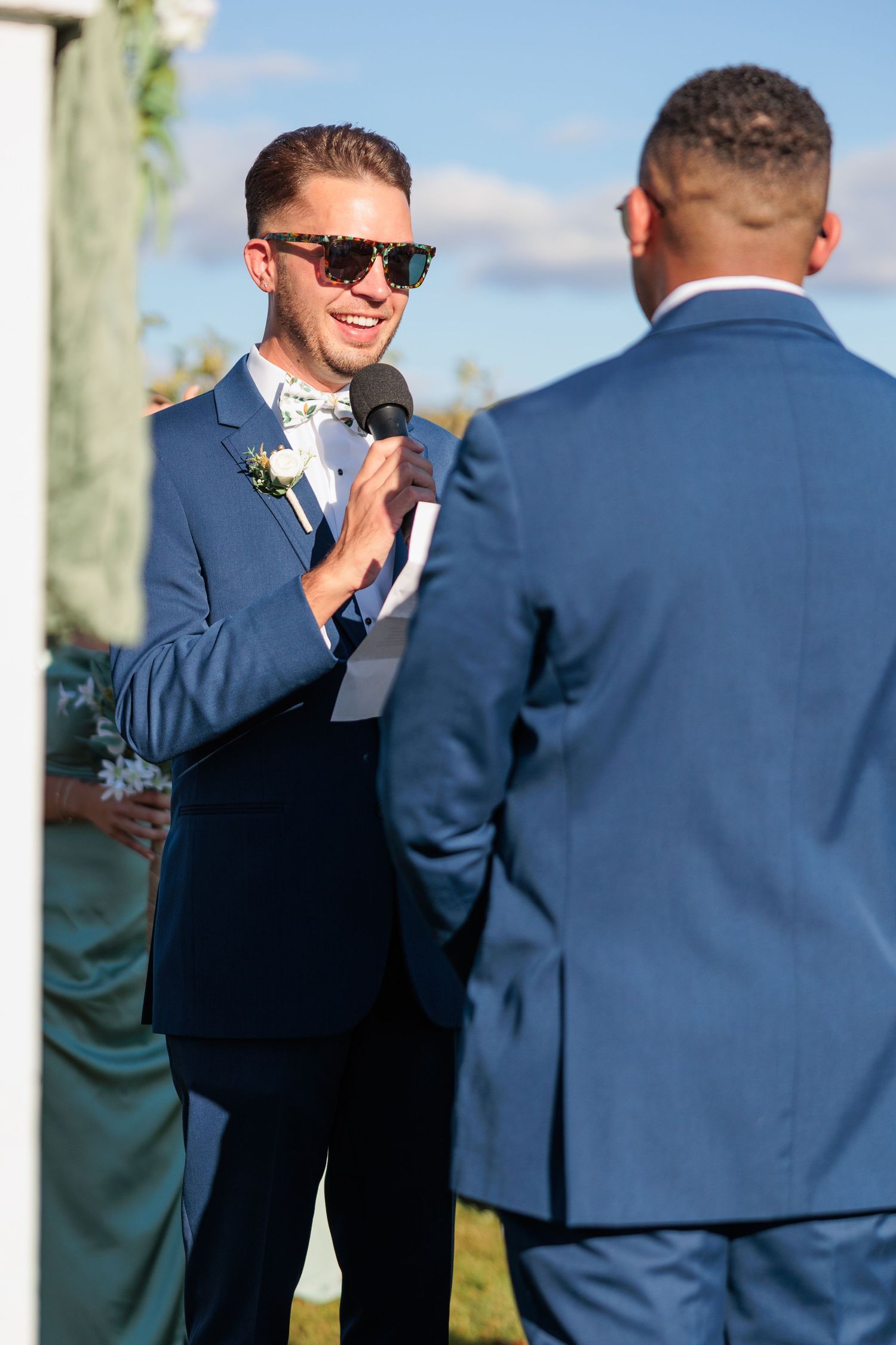 Wedding party members in navy suits interact during outdoor ceremony.