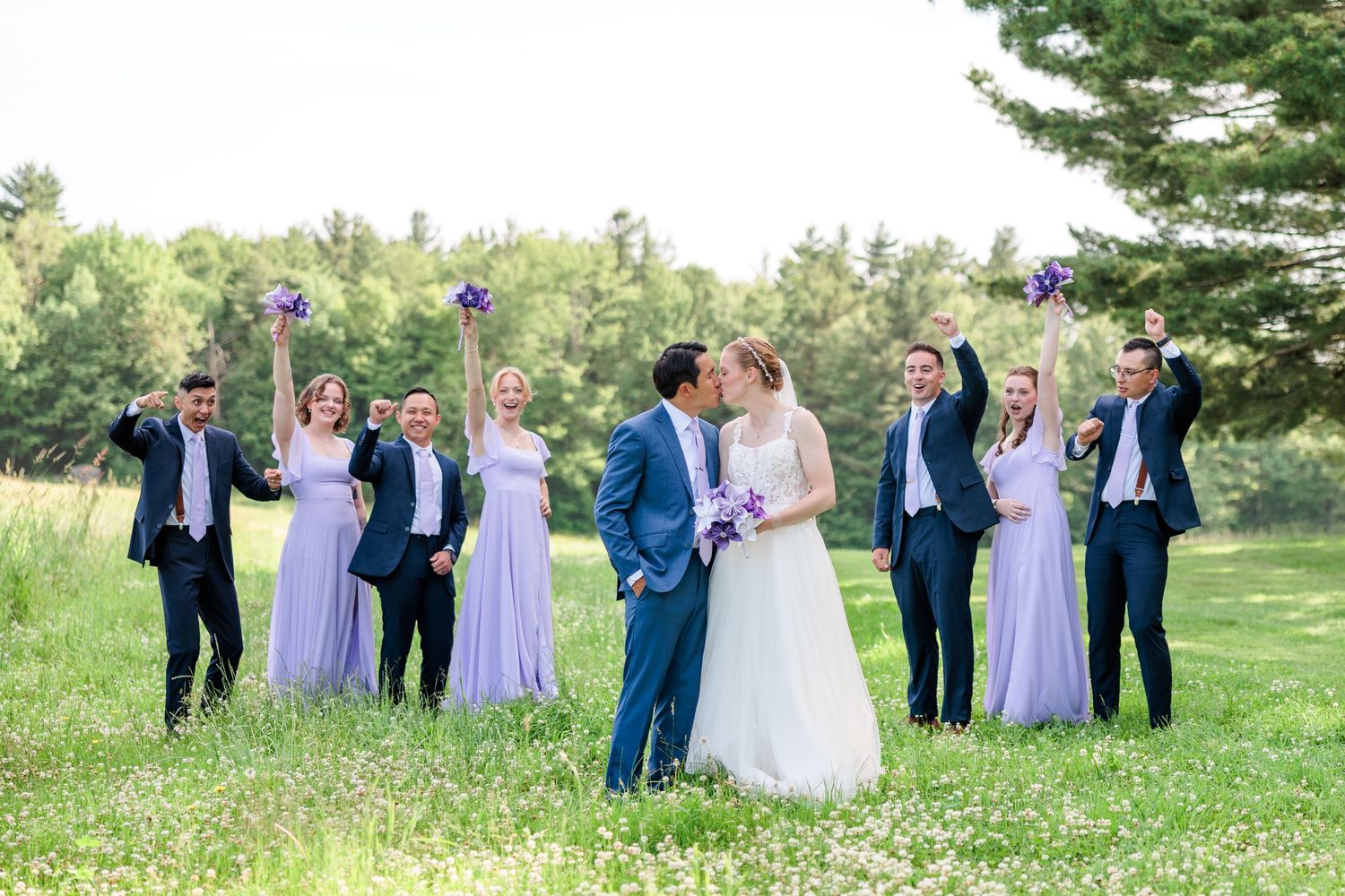 Wedding party in navy suits and lavender dresses celebrating as bride and groom kiss in a green field.