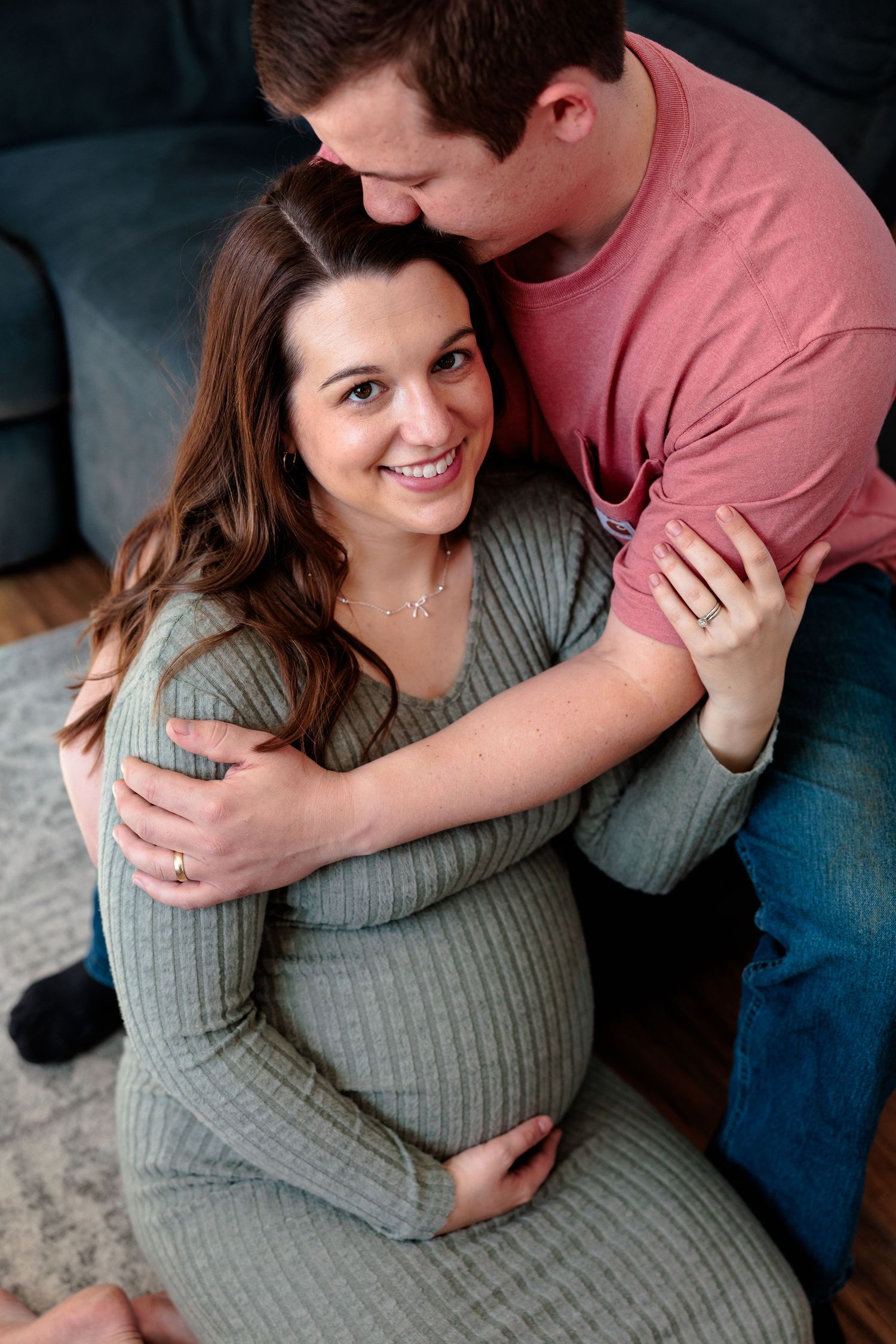A smiling pregnant woman in a green ribbed dress being embraced by her partner while seated on the floor.