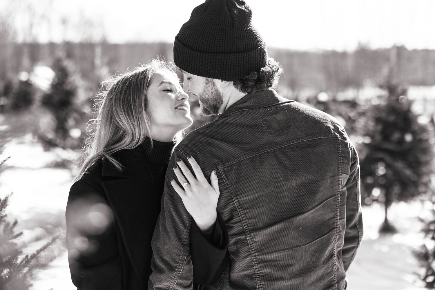 Black and white photo of a laughing couple embracing warmly among snow-covered Christmas trees.
