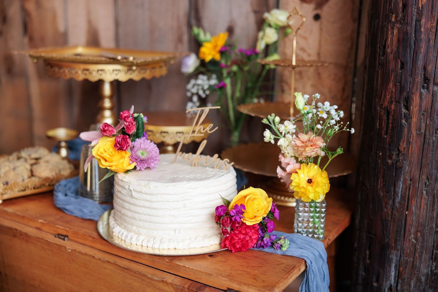 Single-tier white wedding cake decorated with bright flowers on a rustic wooden table in a barn setting.