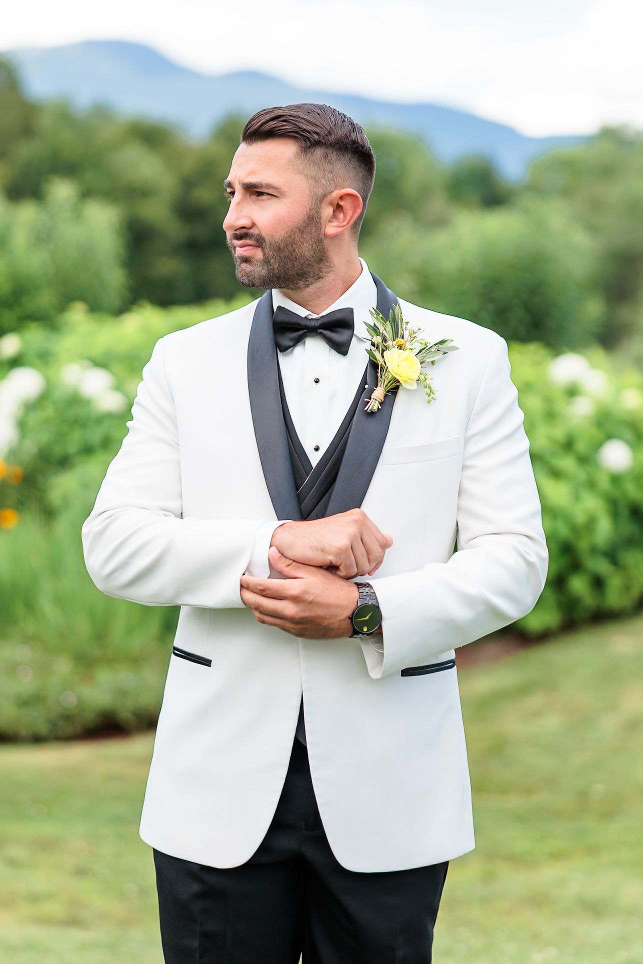 Handsome groom in white tuxedo jacket with black lapels and yellow boutonniere posing outdoors.
