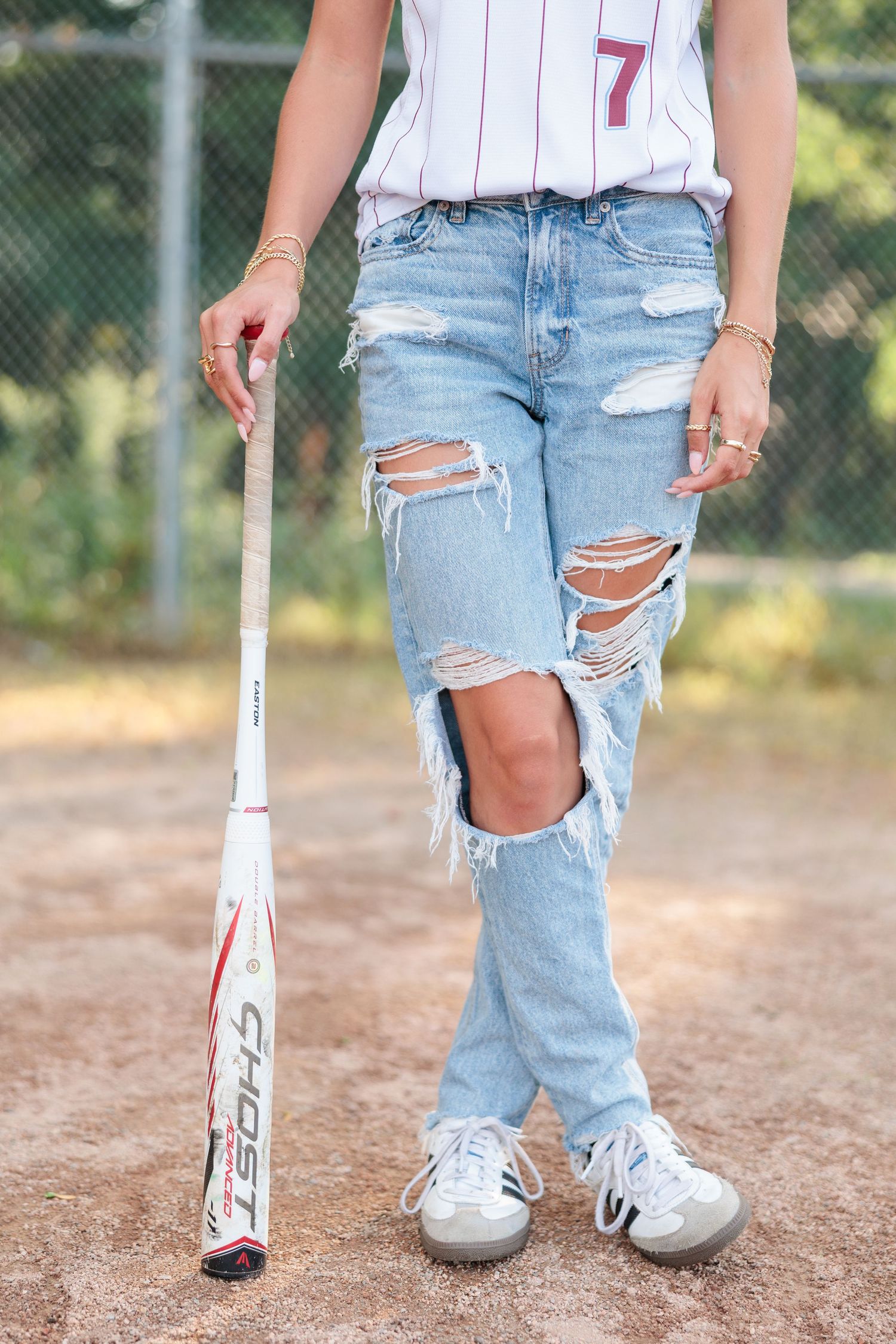Young woman in ripped jeans and baseball jersey holding a Ghost softball bat on a dirt field.