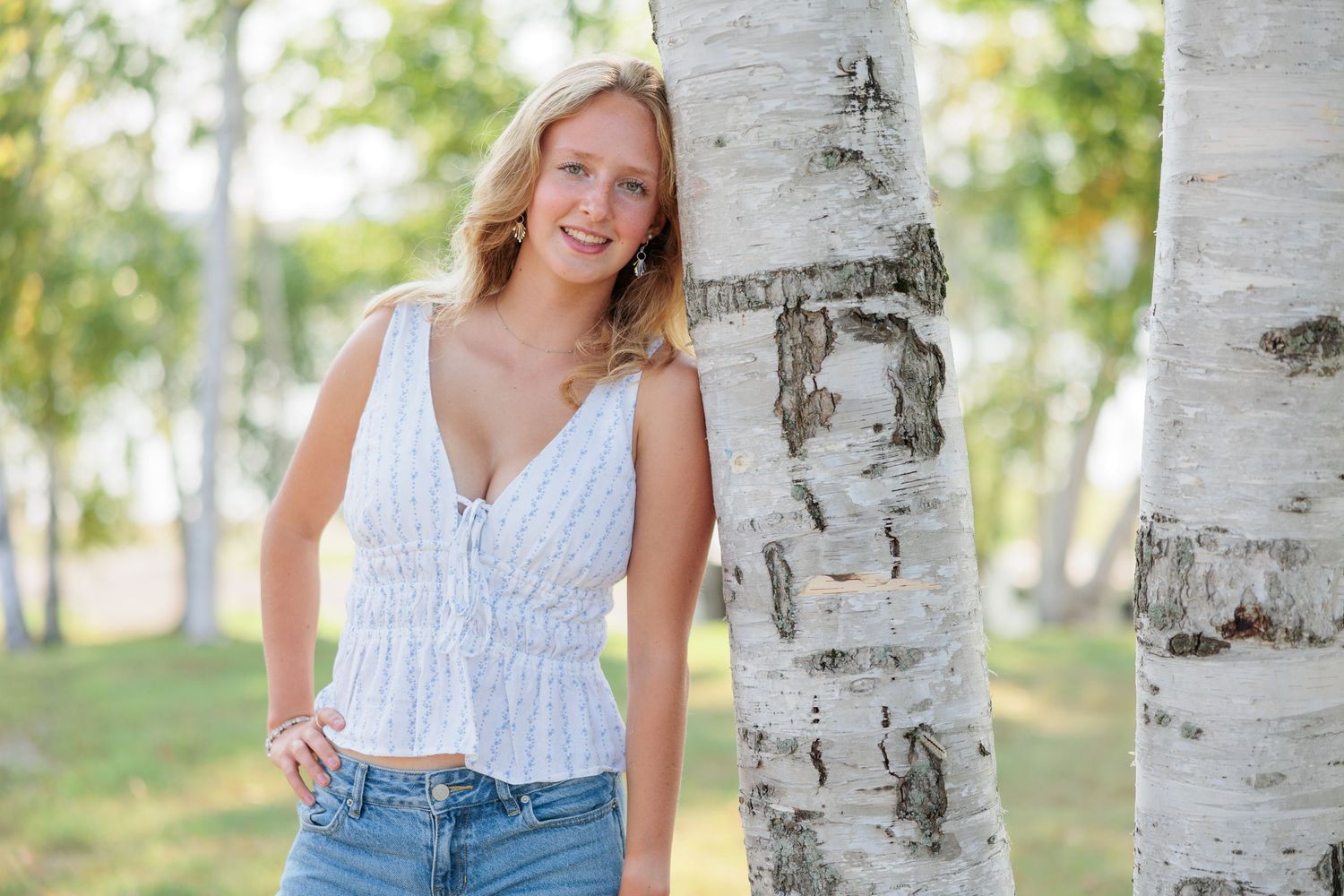 Young blonde woman smiling, leaning against a birch tree in a white top and jeans outdoors.