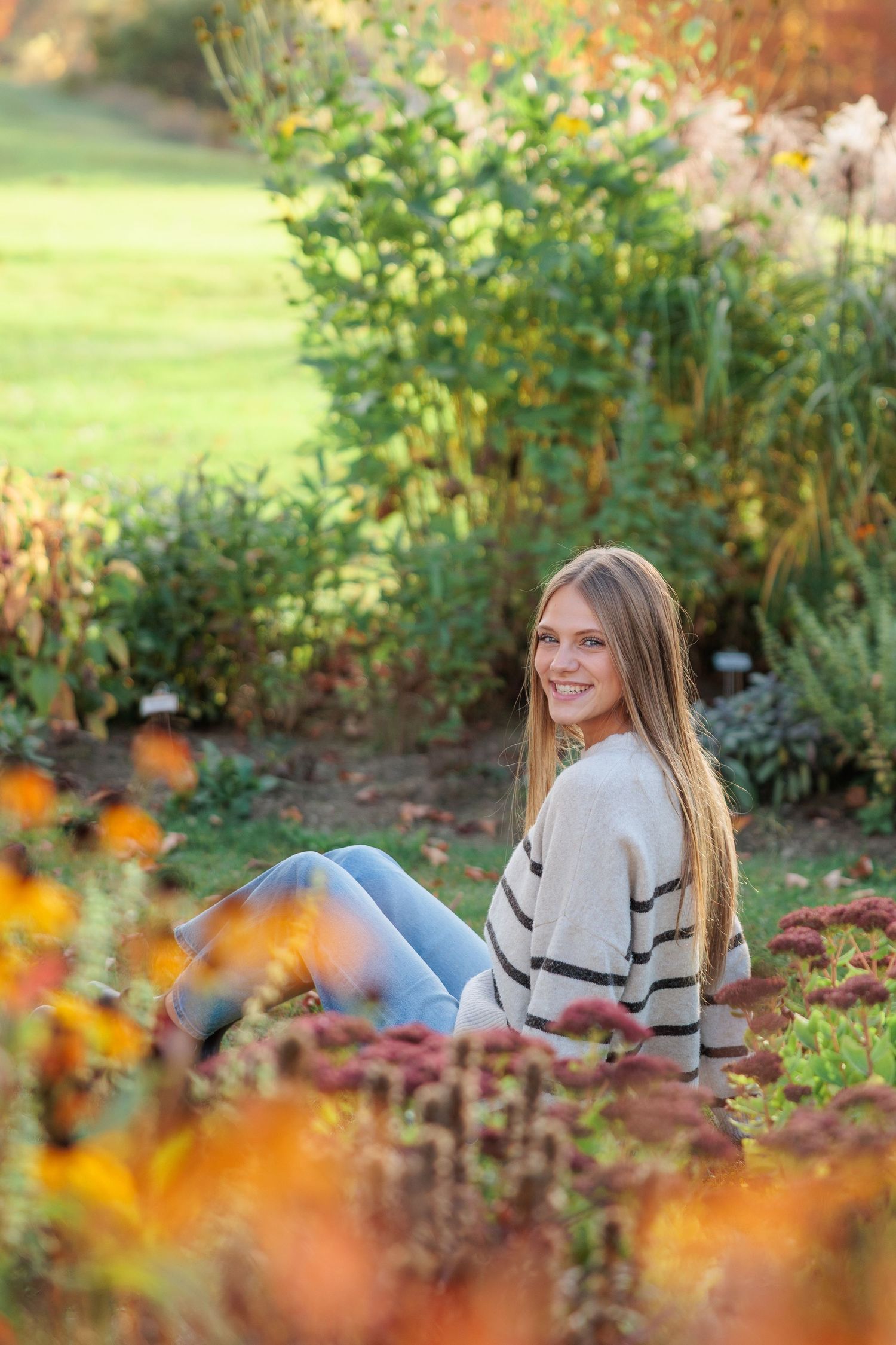 Young woman with long hair smiling in autumn garden, wearing striped sweater and jeans among colorful fall flowers.