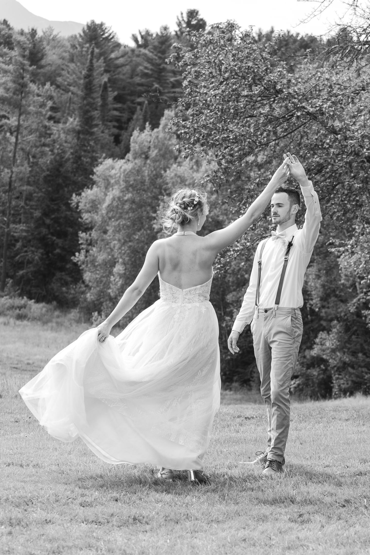 Black and white photo of groom spinning bride in a flowing dress outdoors in a scenic meadow.