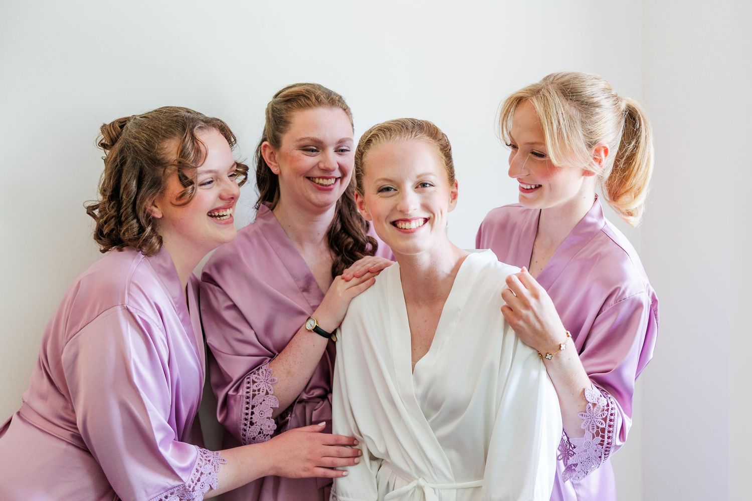 Smiling bride in white robe surrounded by three bridesmaids in lavender robes during wedding morning preparations.