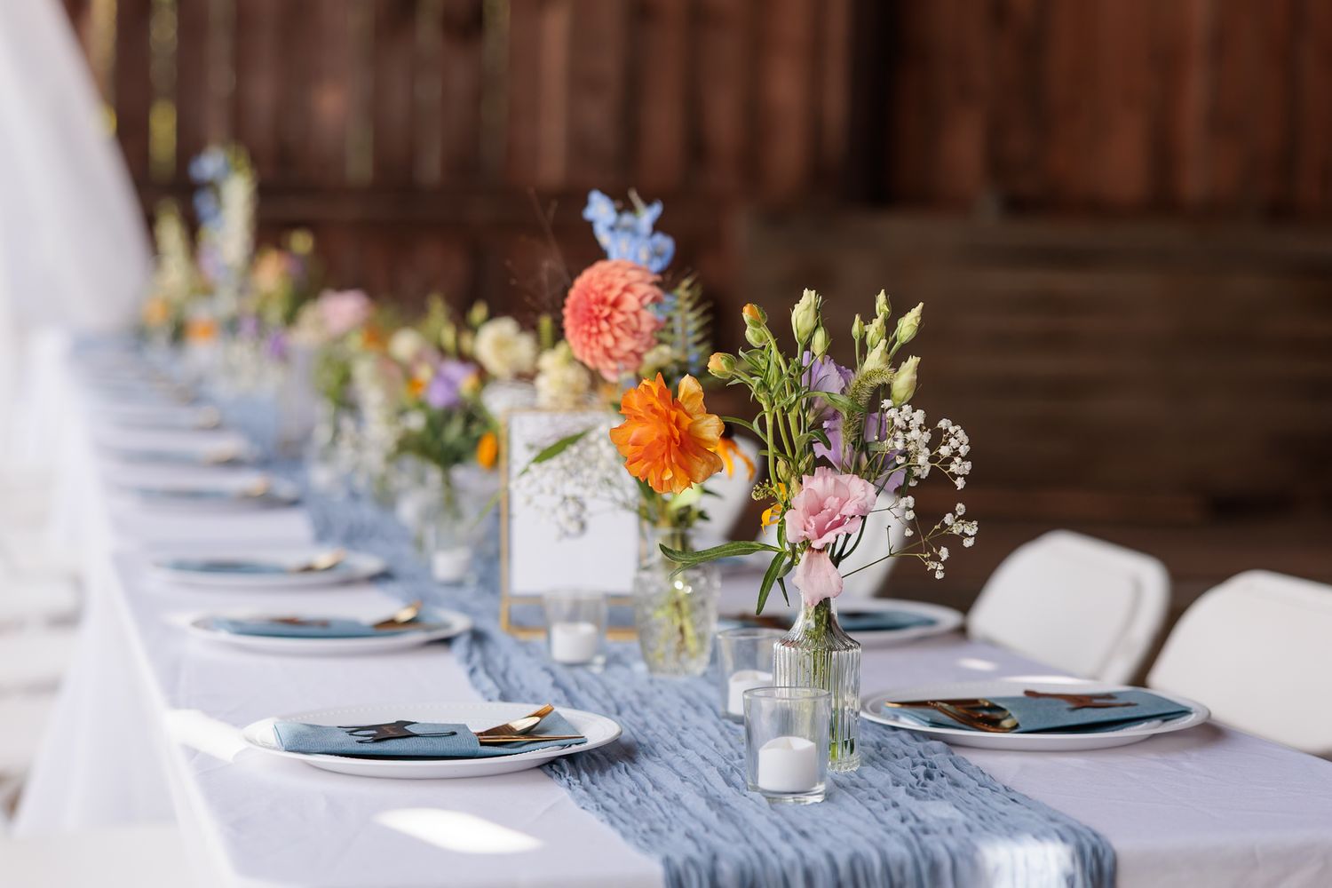 Long rustic barn wedding reception table with blue cheesecloth runner, vibrant wildflower arrangements, and gold cutlery.