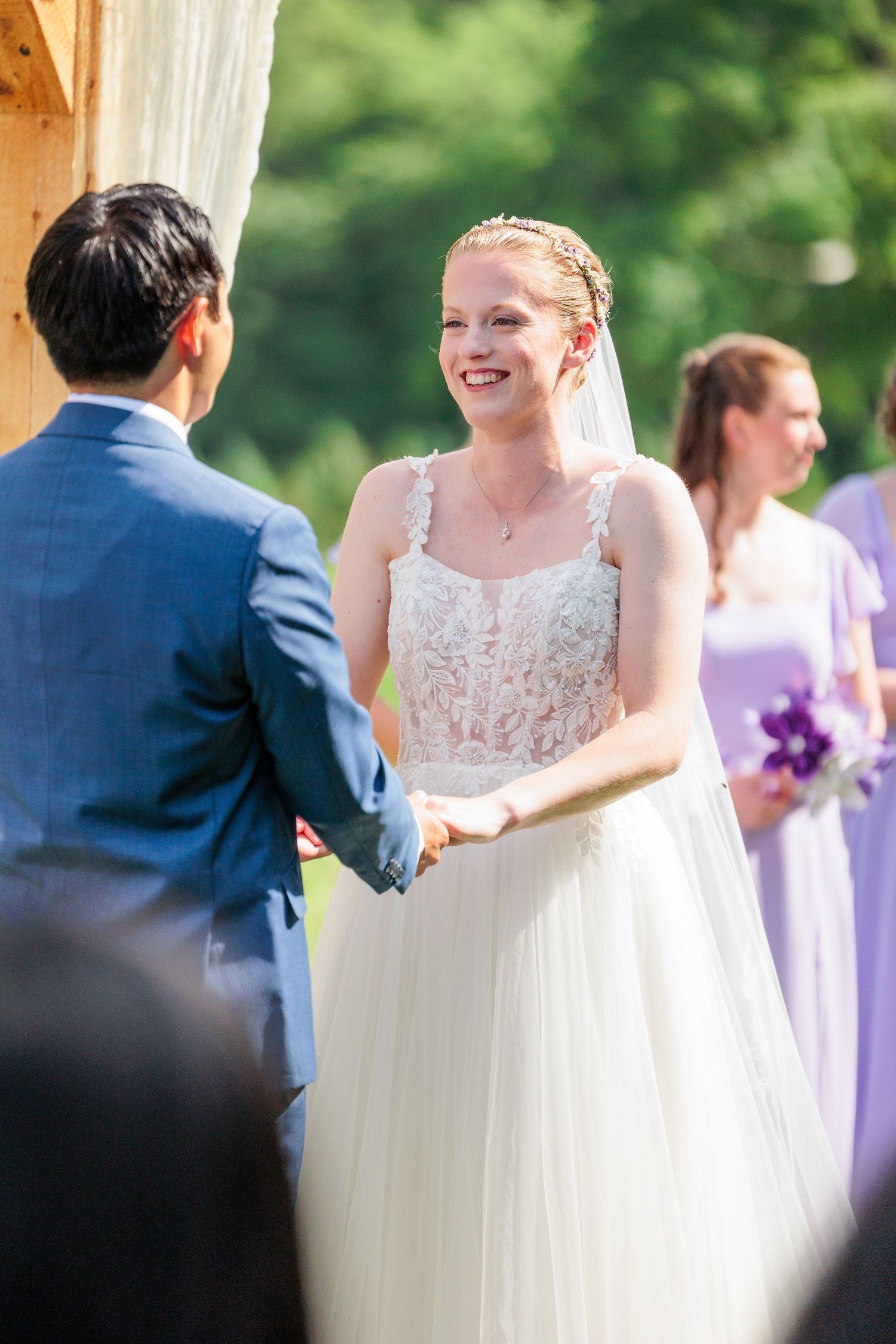 Radiant bride in lace gown smiling while holding hands with groom during outdoor wedding ceremony.