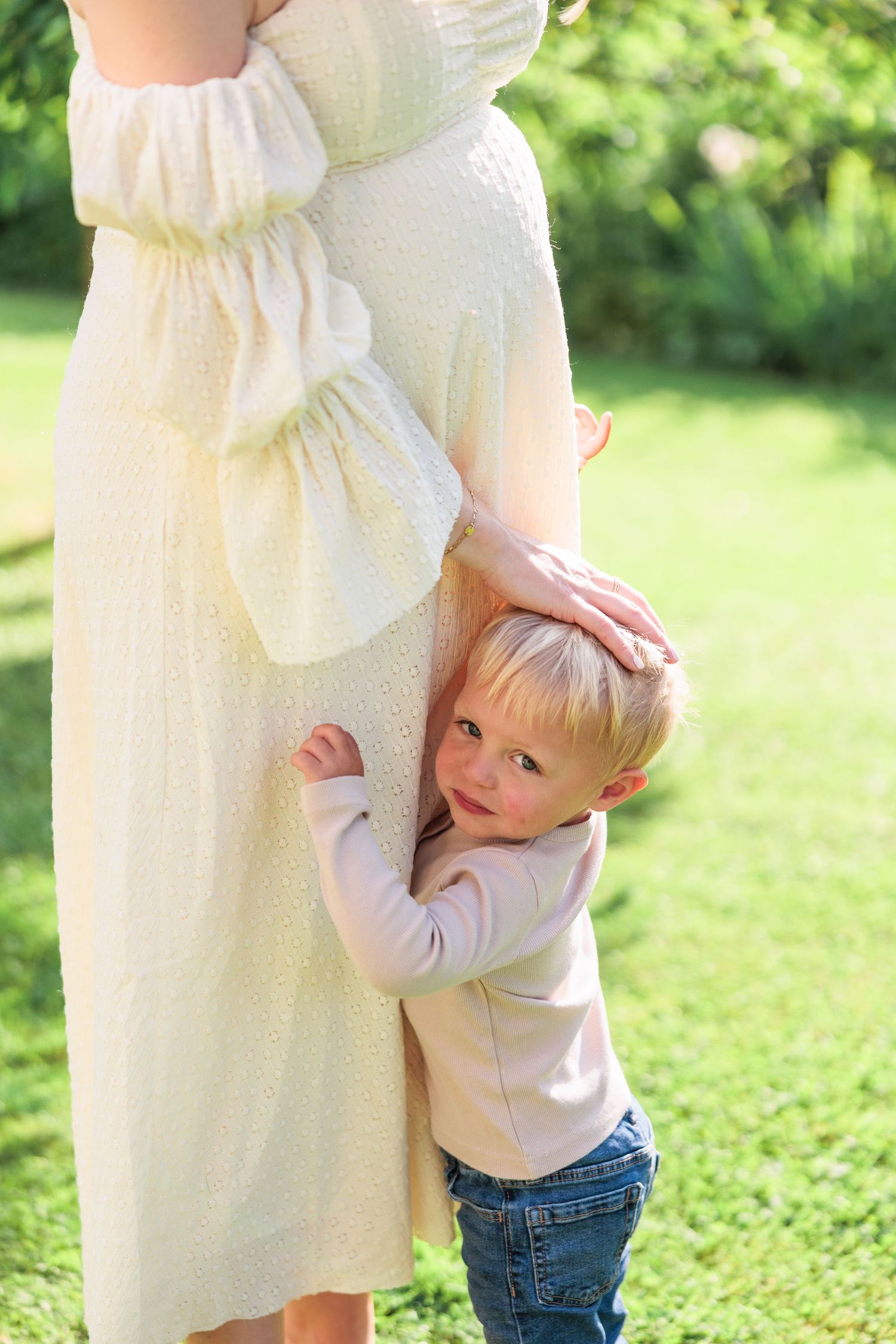 Toddler hugging pregnant mothers belly in a sunny green garden during maternity session.