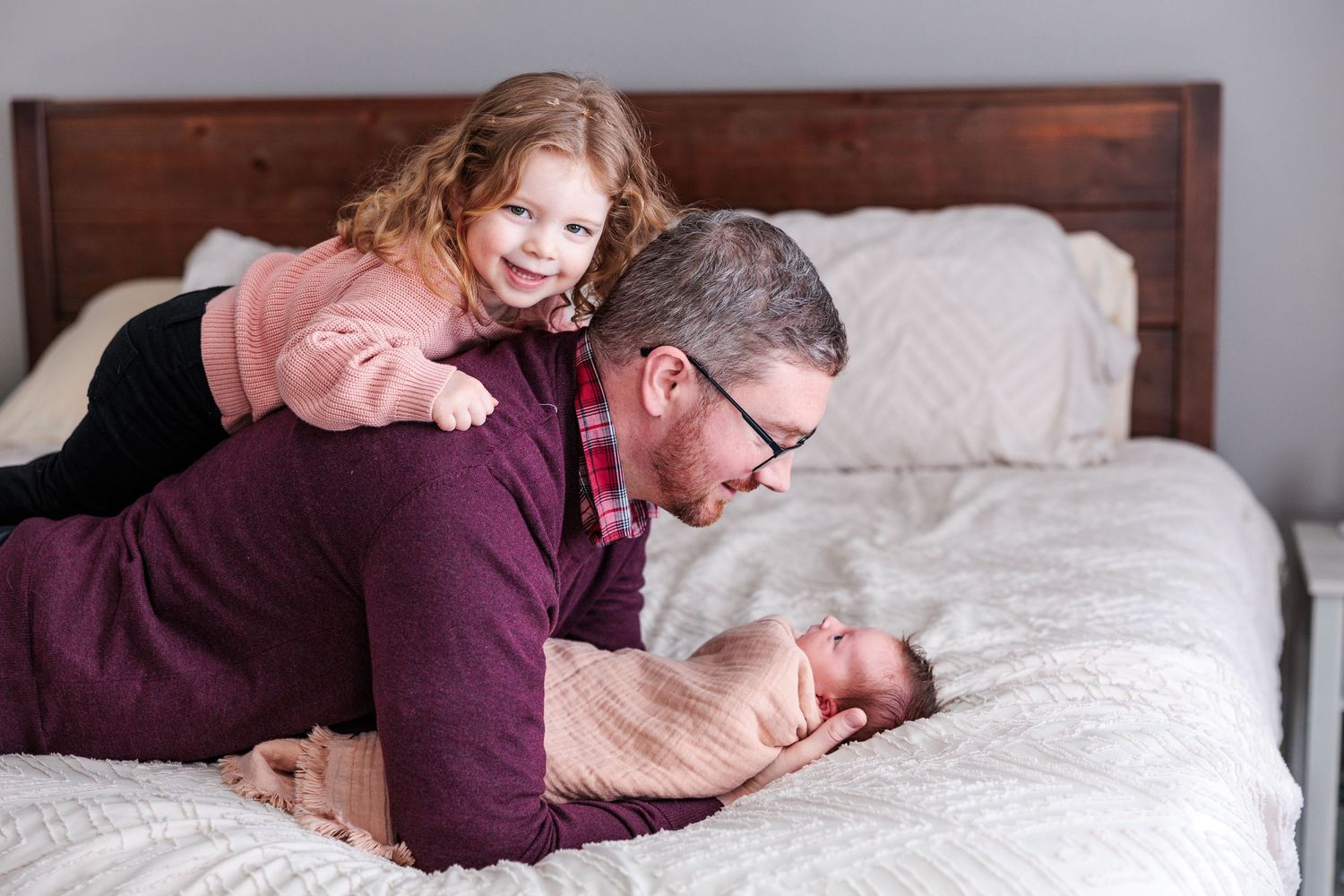 Father lying on bed with toddler daughter on his back while looking at newborn baby.