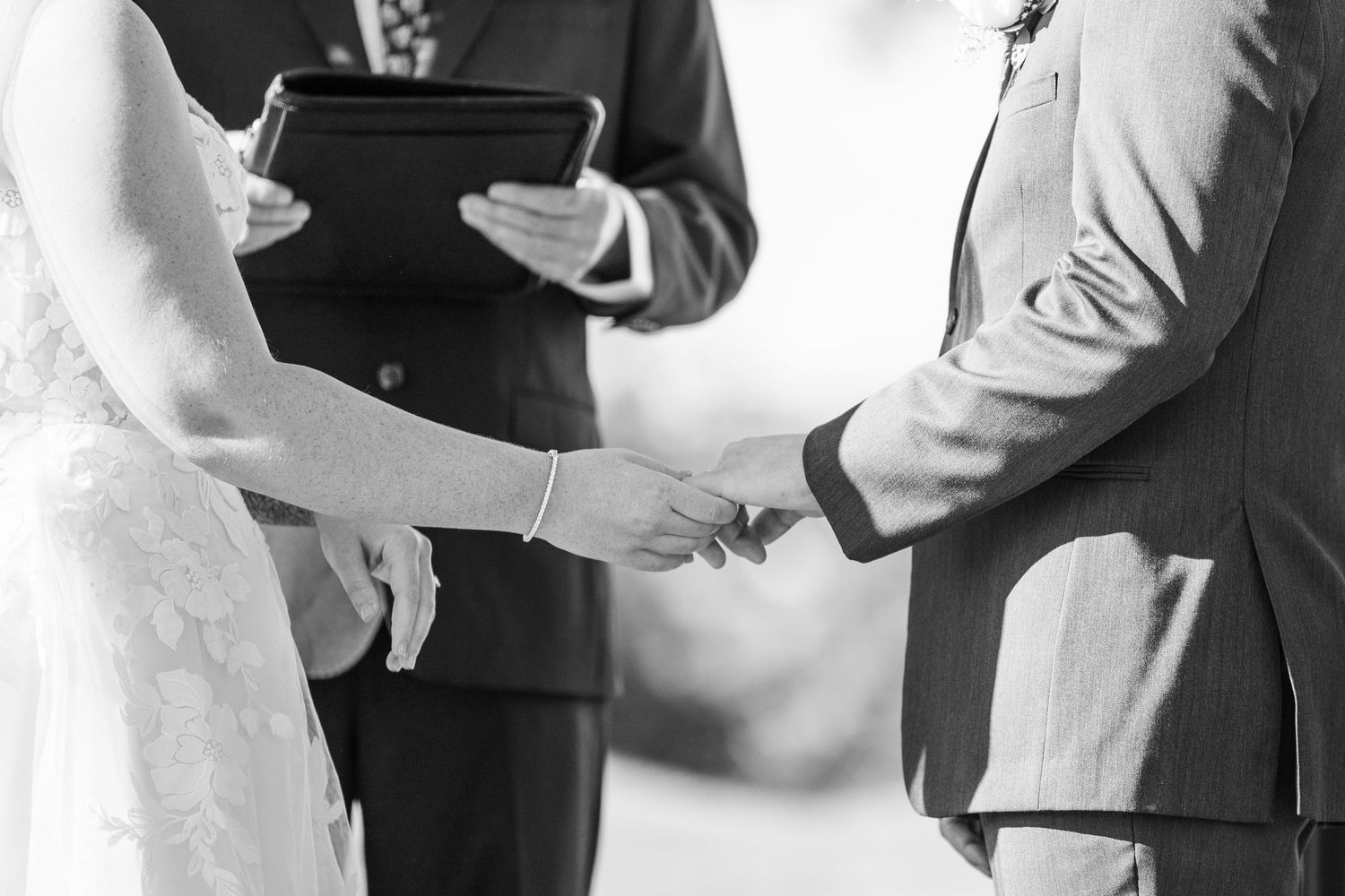 Black and white close-up of bride and groom exchanging rings during outdoor wedding ceremony.