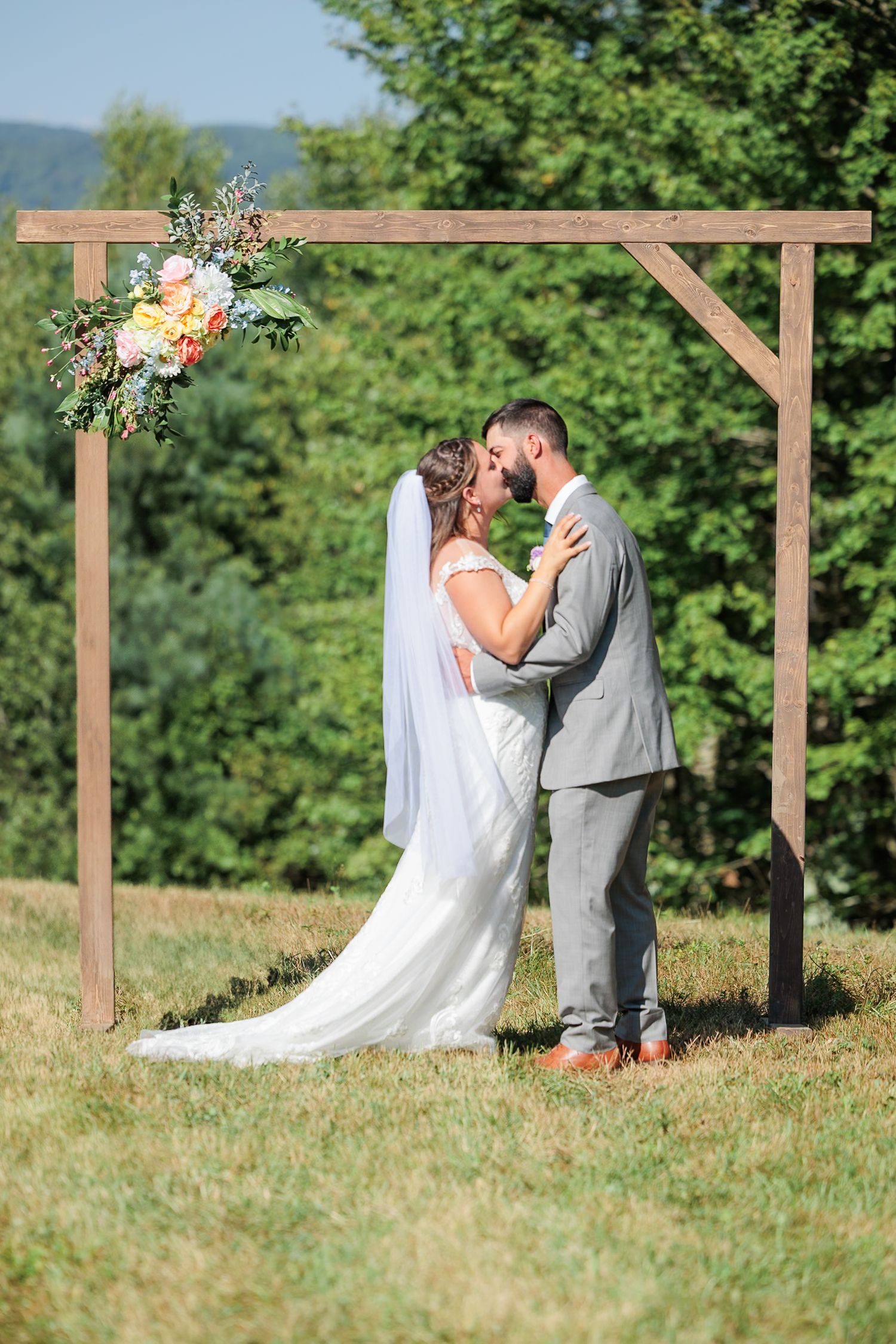 Bride and groom share first kiss under rustic wooden arch adorned with colorful flowers in open field.
