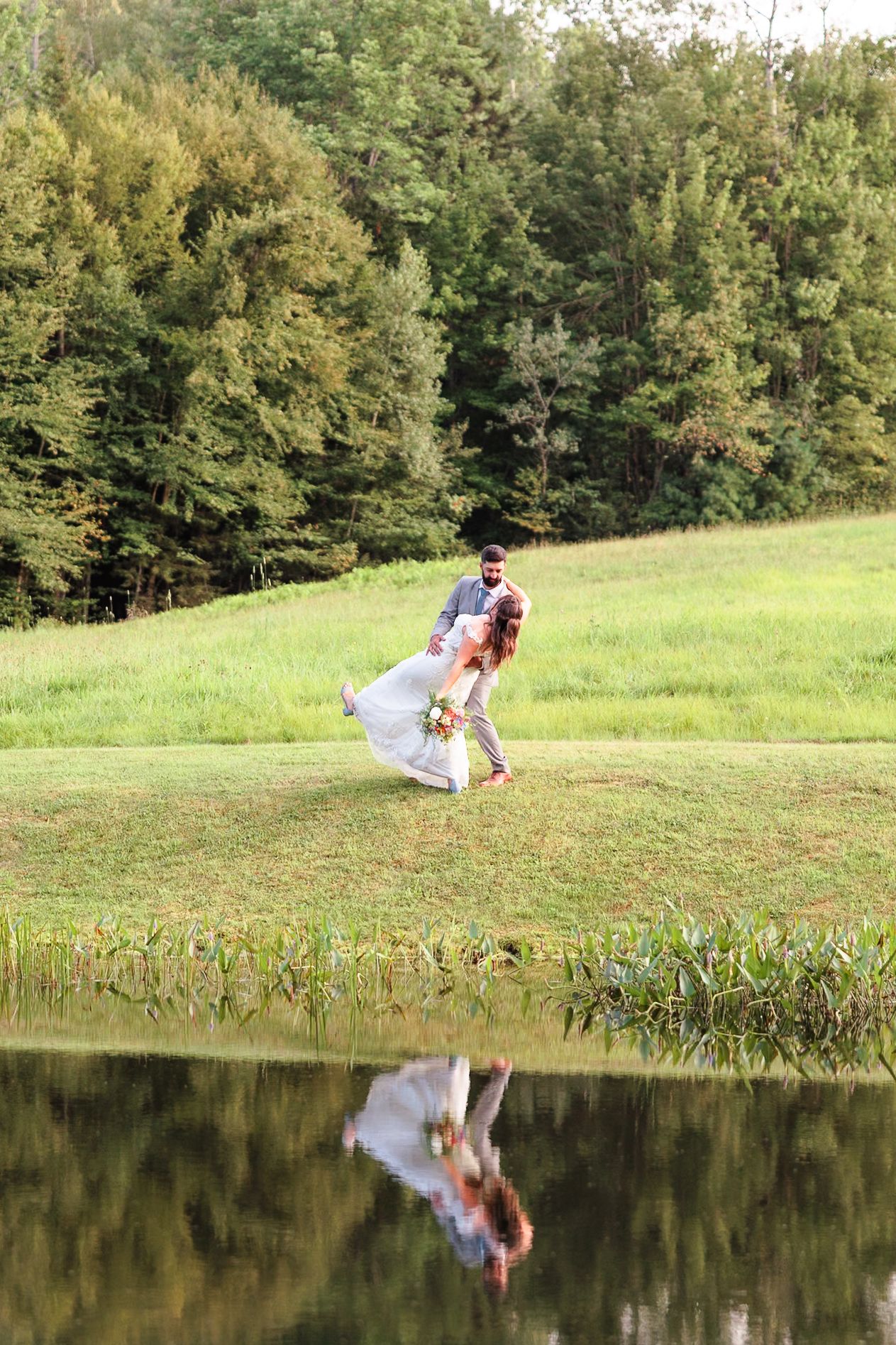 Groom dipping bride in white dress near a reflective pond surrounded by green trees.