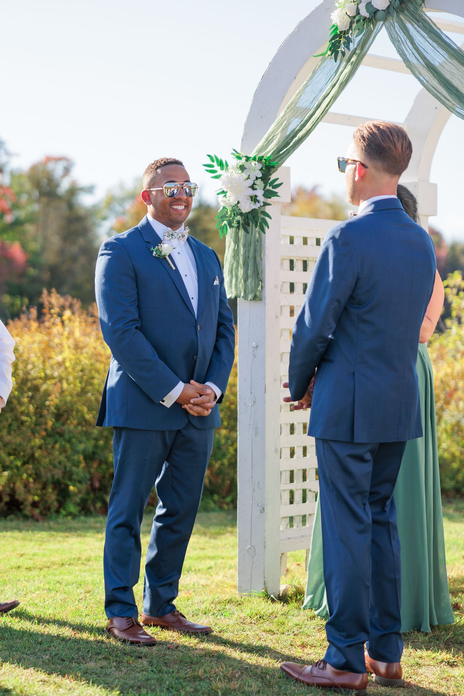 Wedding party members in navy suits stand by a white lattice arch decorated with sage green fabric.