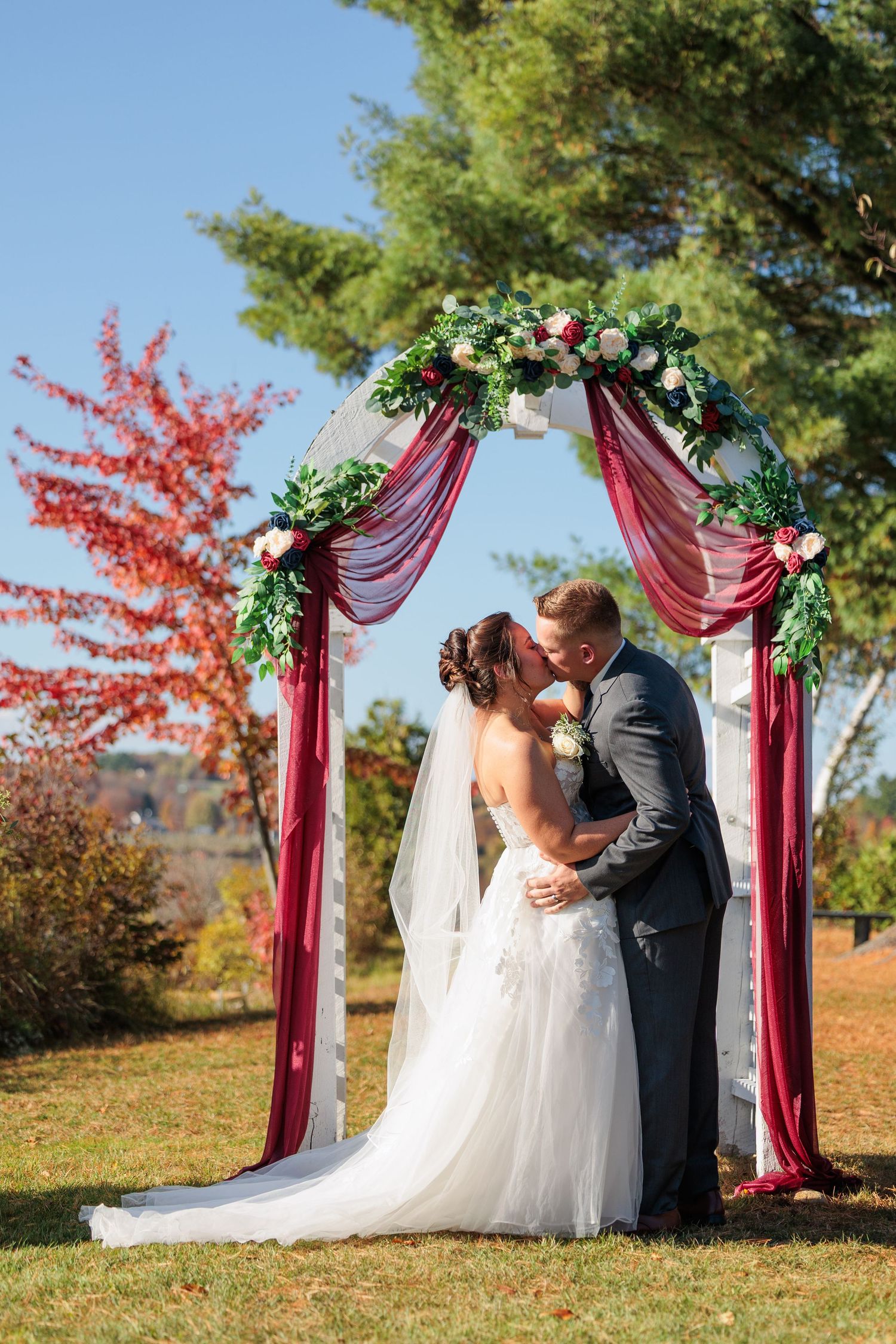 Romantic outdoor autumn wedding ceremony under burgundy draped arch with greenery and fall foliage in background.