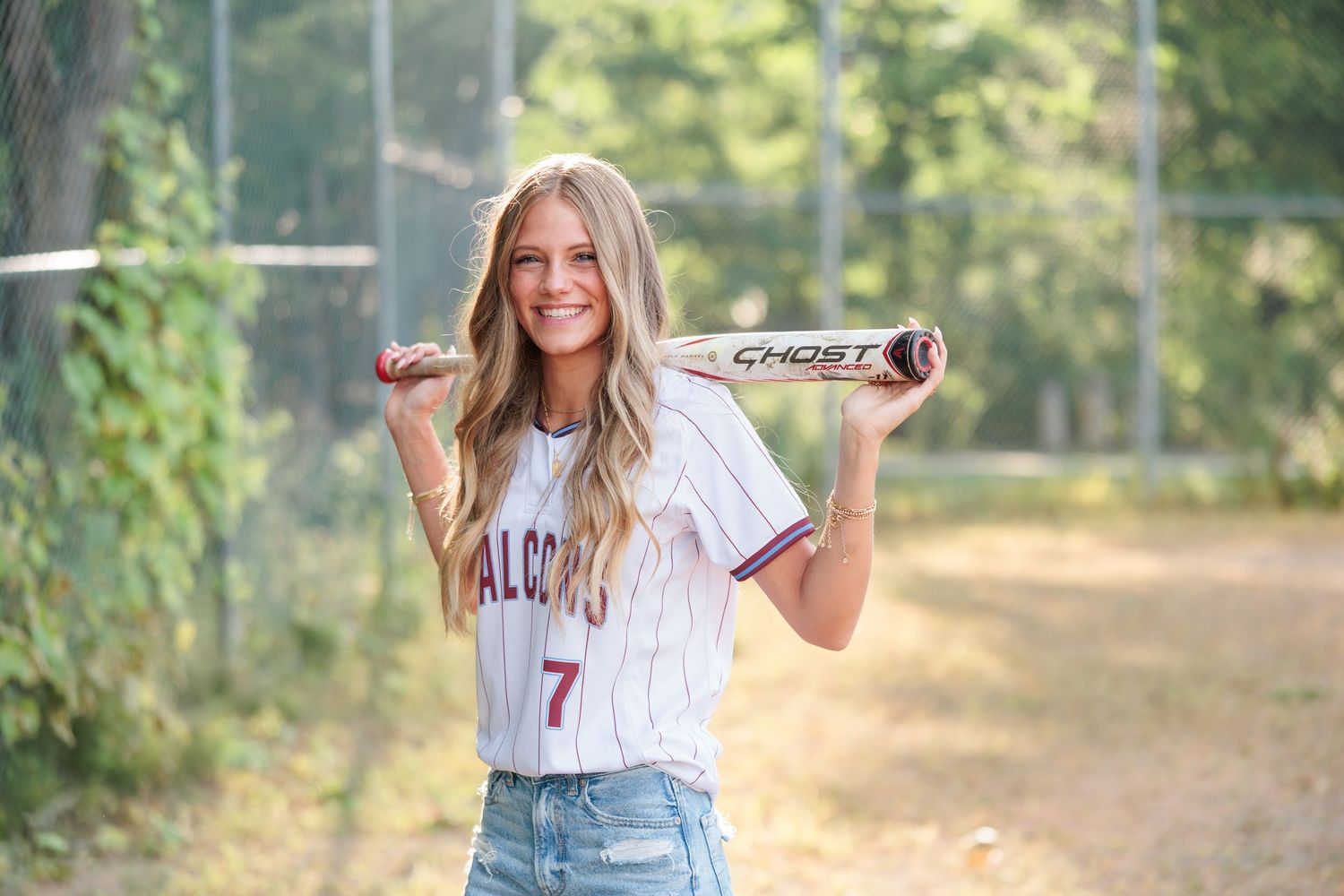 Young female softball player in Falcons jersey number 7 holding a Ghost bat behind her shoulders on a field.