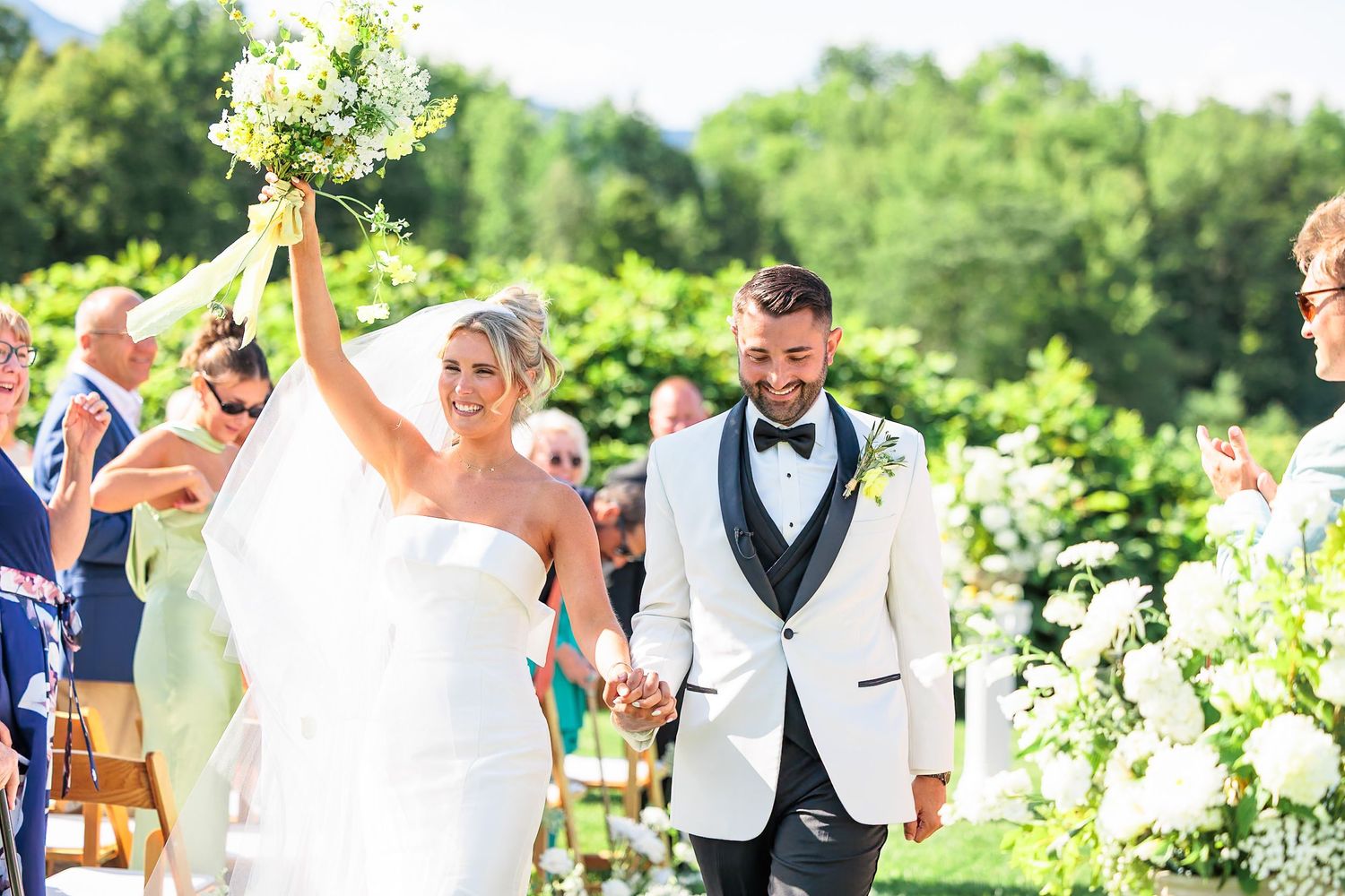 Joyful bride raises bouquet while walking down the aisle with groom after outdoor wedding ceremony.