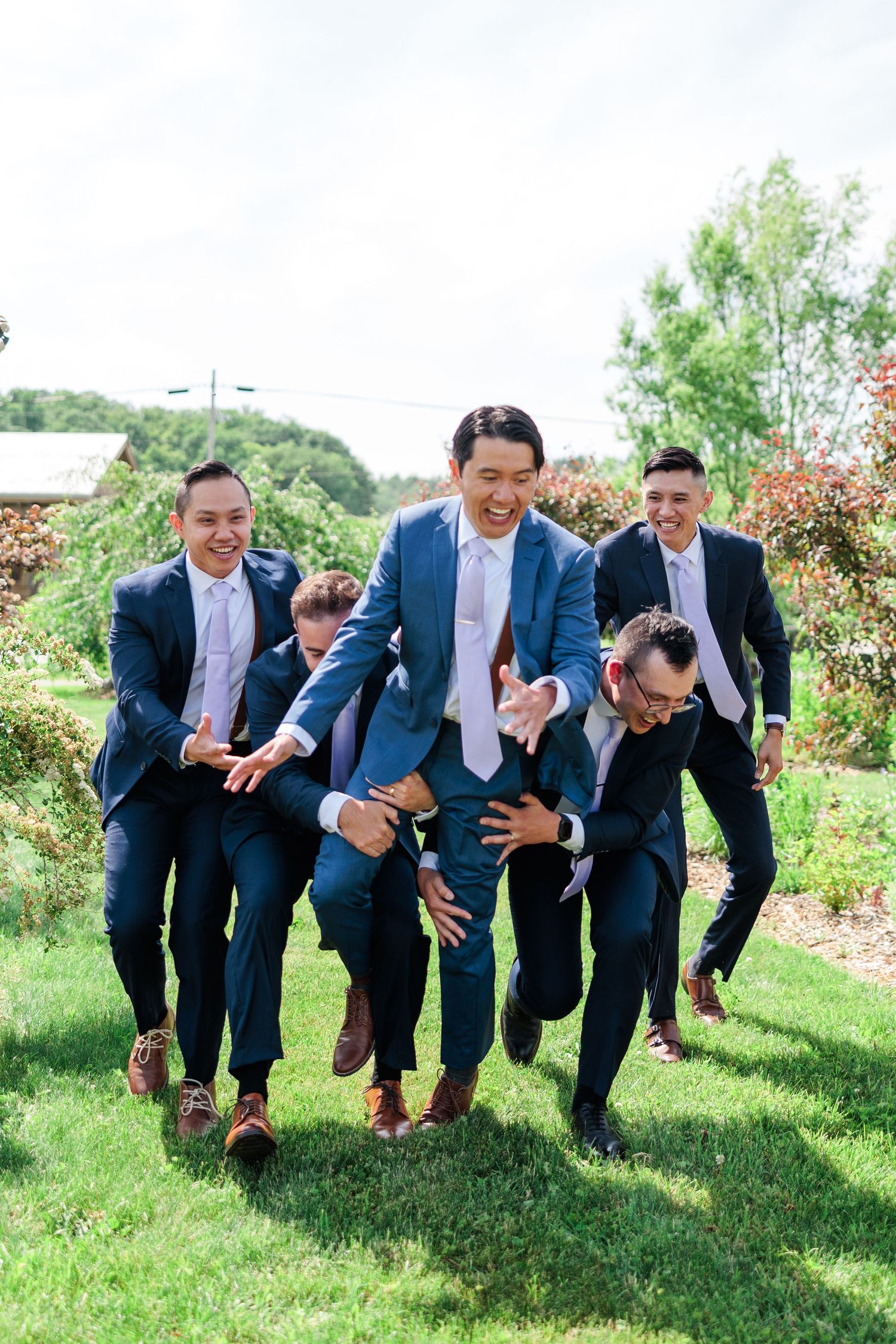Groomsmen in navy suits laughing and playing around outdoors on green grass at a wedding celebration.