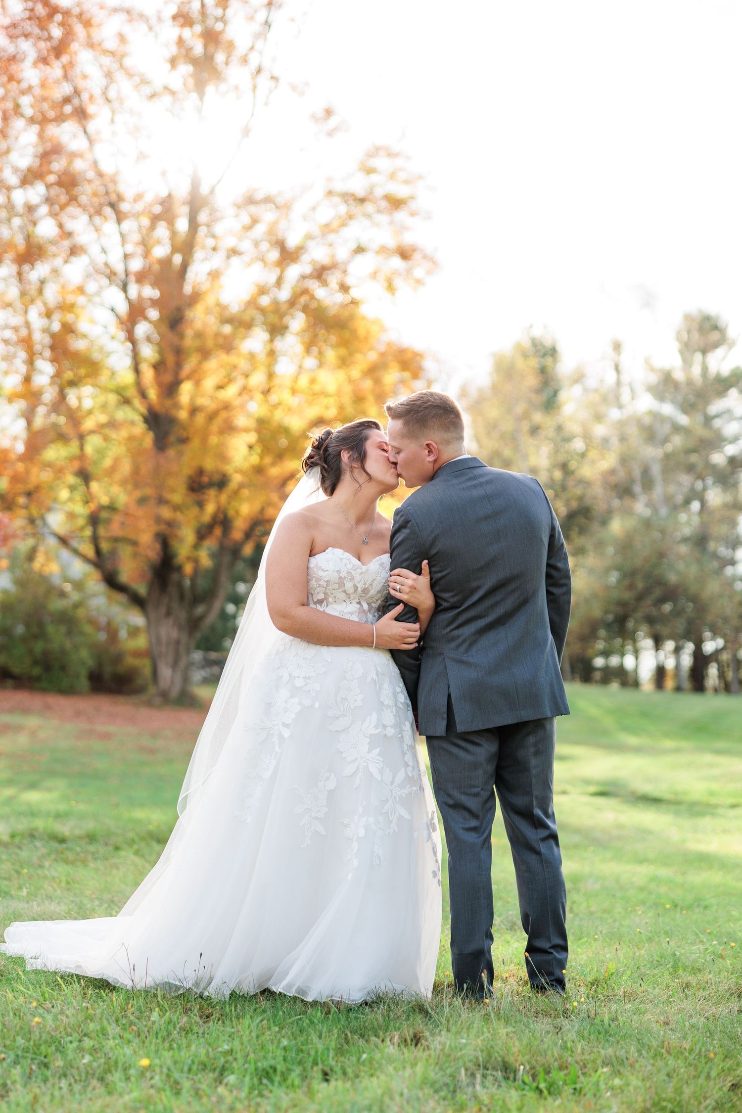 Bride and groom sharing a kiss outdoors surrounded by colorful fall trees at sunset.