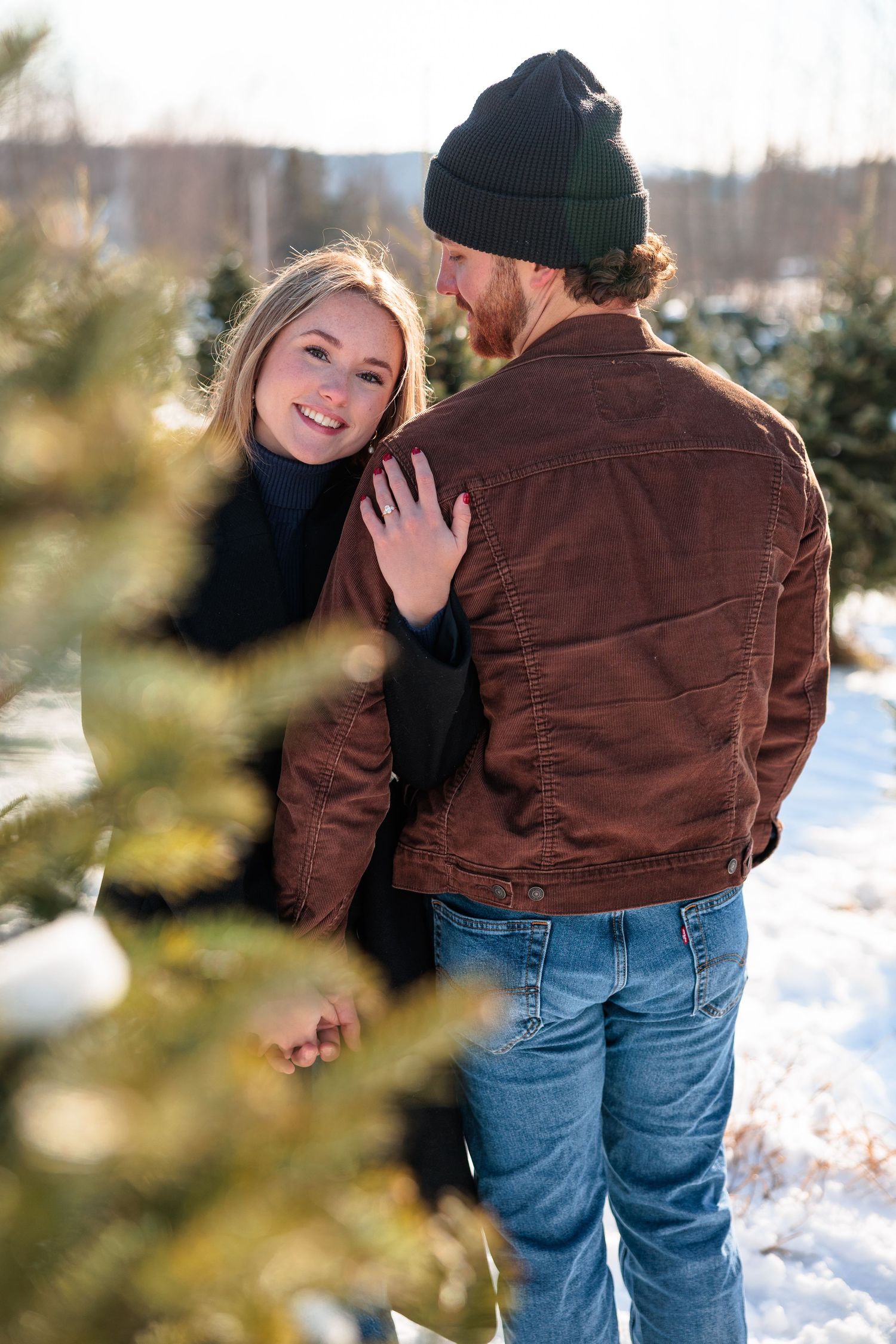 Smiling woman rests her hand on her partners brown jacket at a snowy Christmas tree farm.