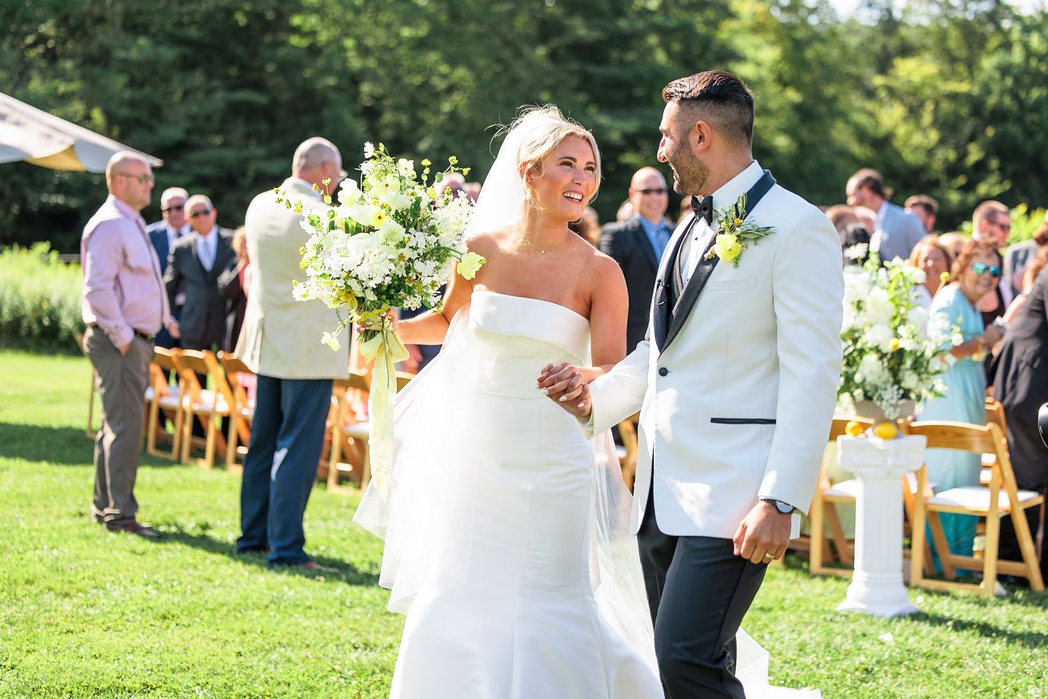 Smiling newlyweds walk hand in hand down aisle after outdoor garden wedding ceremony in sunshine.