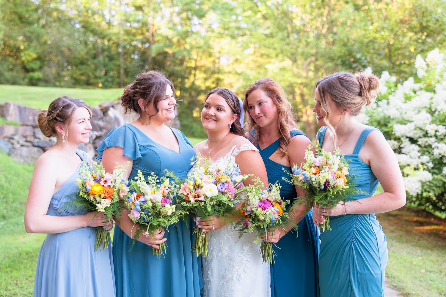 Bride in white lace gown stands with four bridesmaids in blue dresses holding colorful wildflower bouquets.