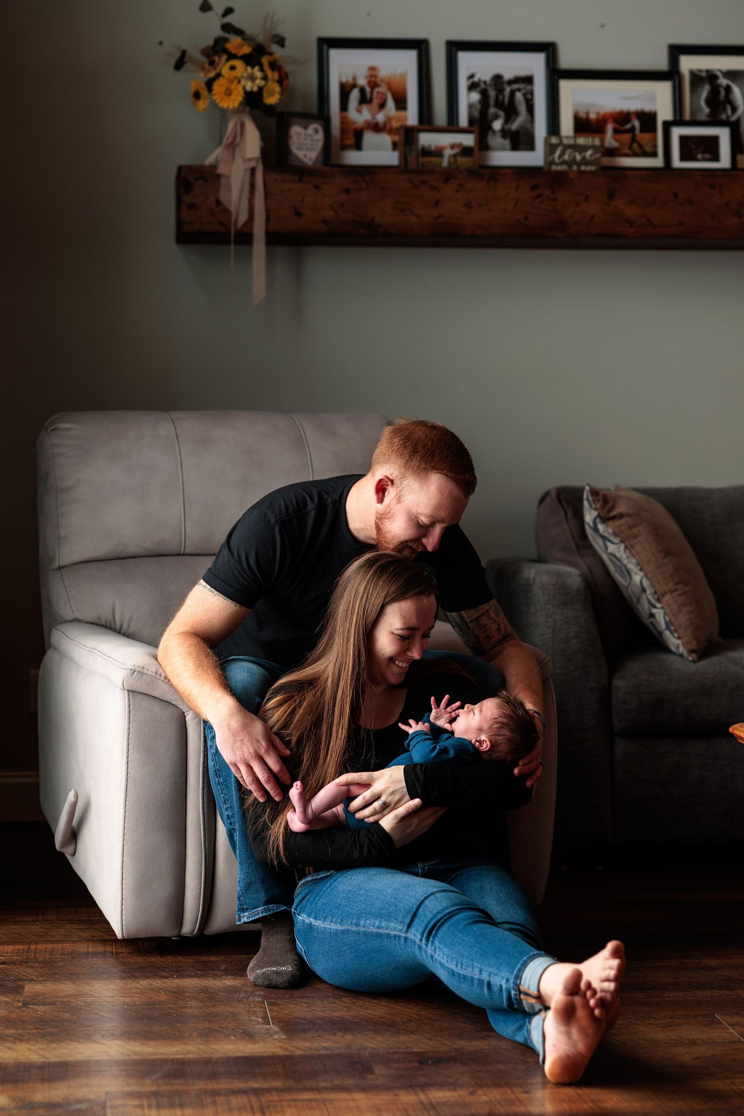 Happy couple sitting on living room floor cuddling their newborn baby near a gray sofa.