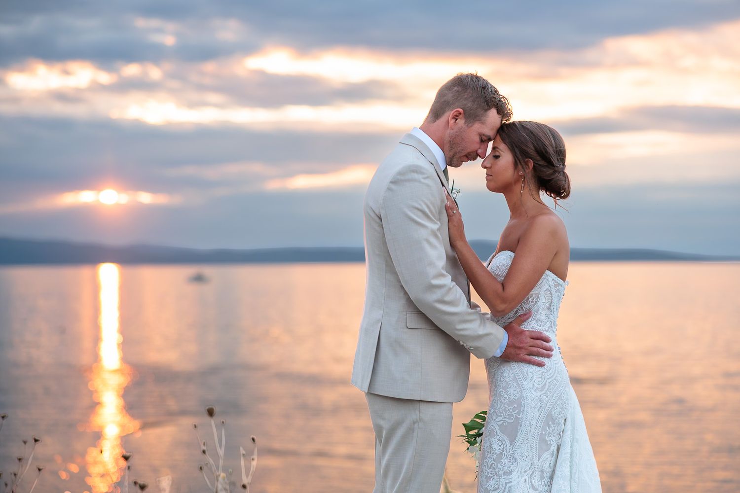 Romantic bride and groom embrace at sunset by the water during golden hour wedding portrait.
