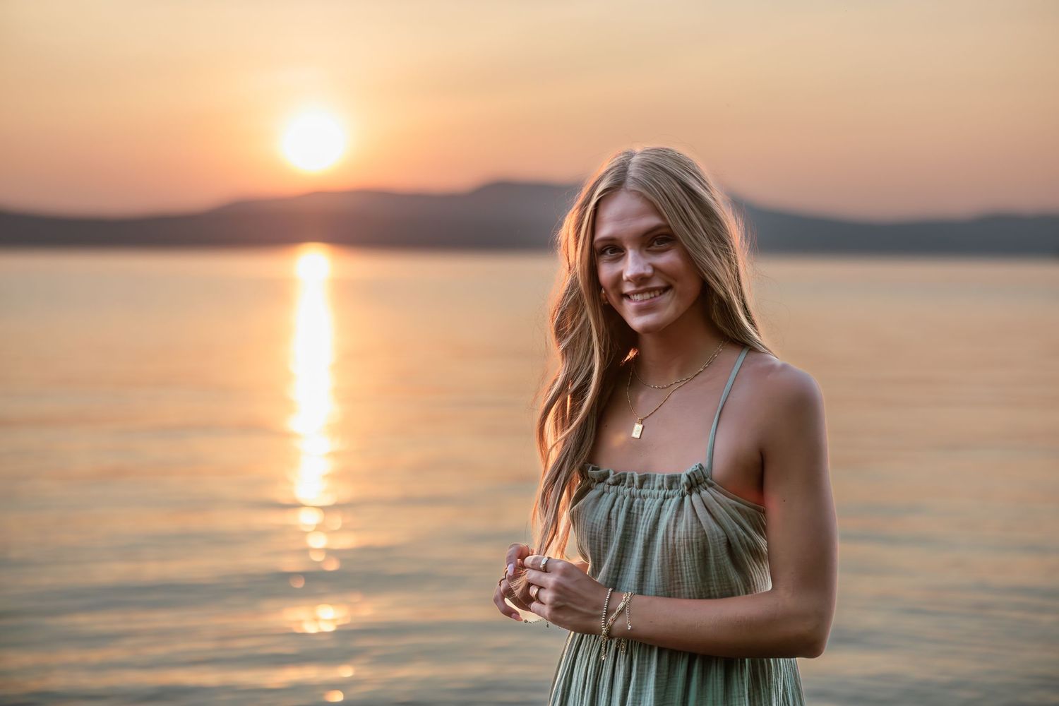 Smiling blonde woman in sage green dress posing by a lake at golden sunset with mountains in background.
