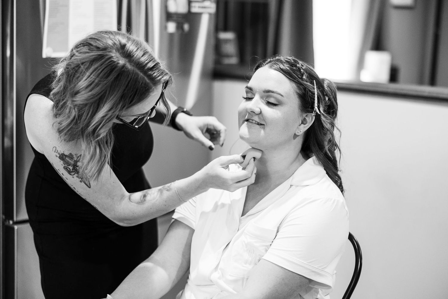 Makeup artist applying lipstick to bride seated in chair during bridal beauty preparations in black and white.