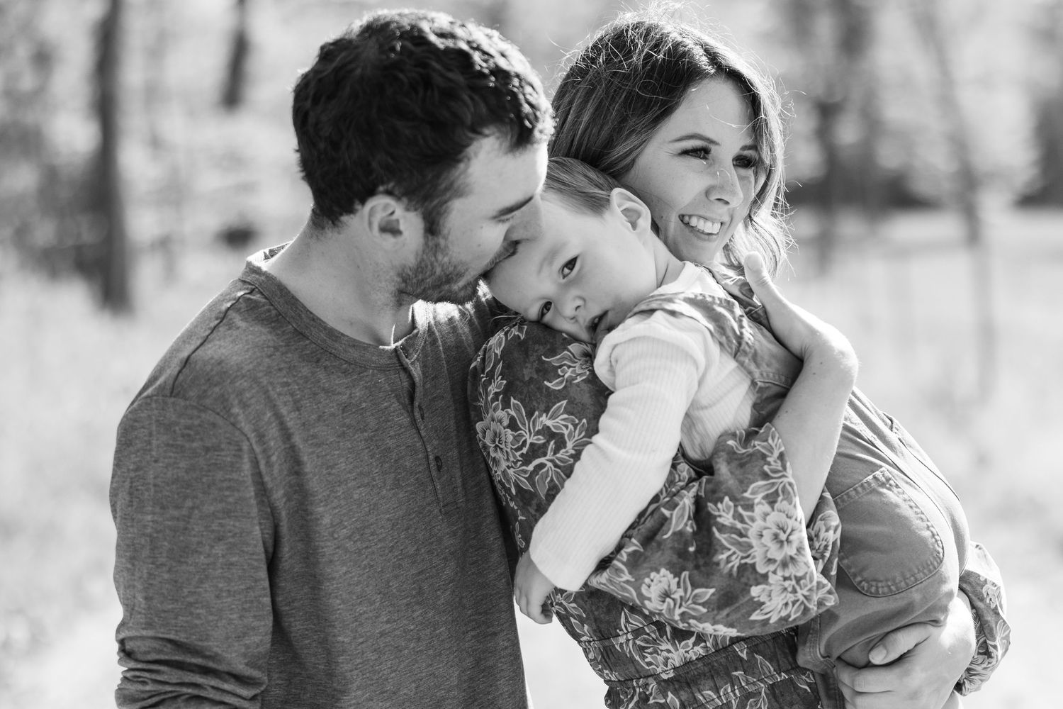 Black and white photo of smiling parents with father kissing young daughter outdoors in nature.