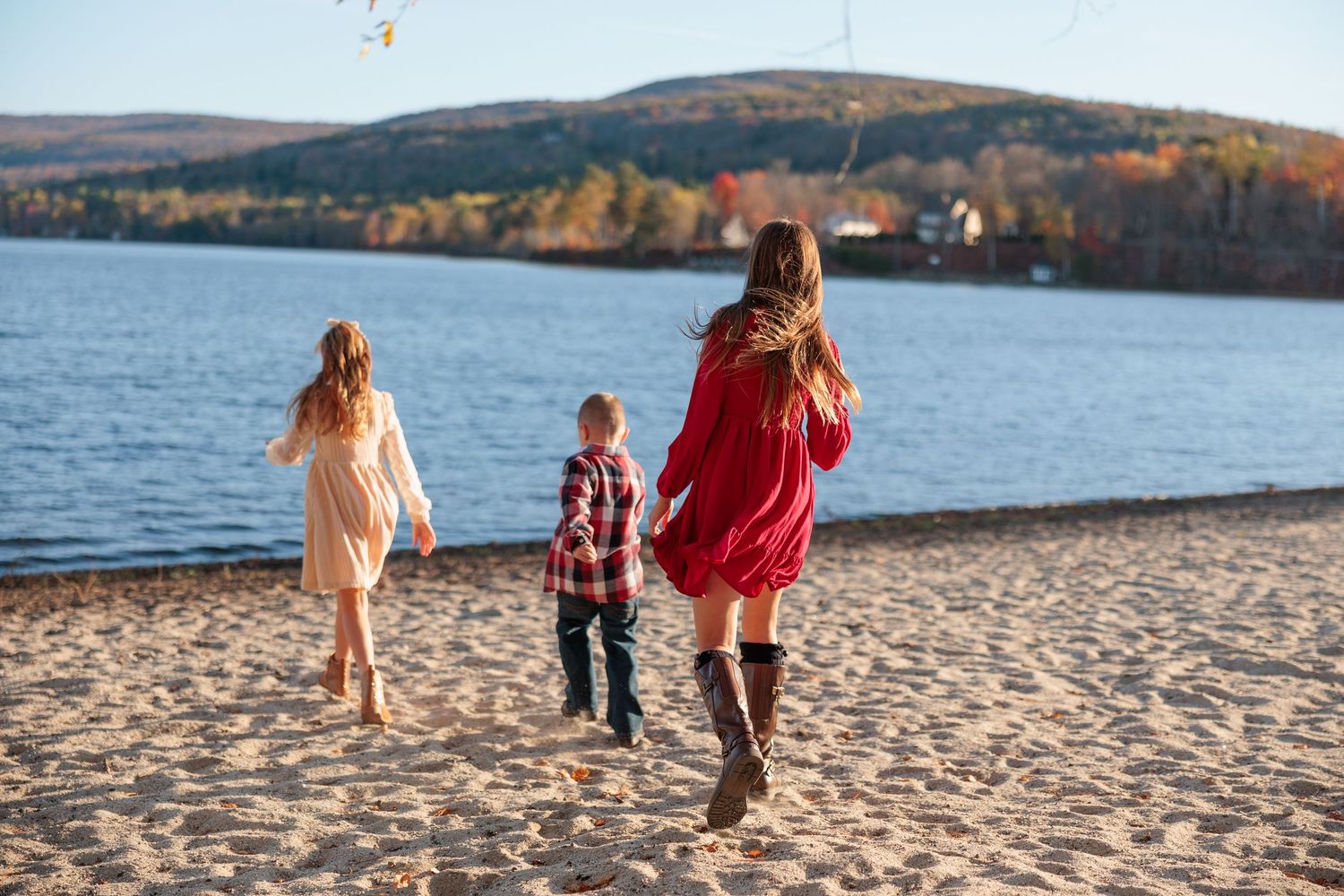 Mother in red dress walking with two children along sandy lakeside beach in autumn setting.