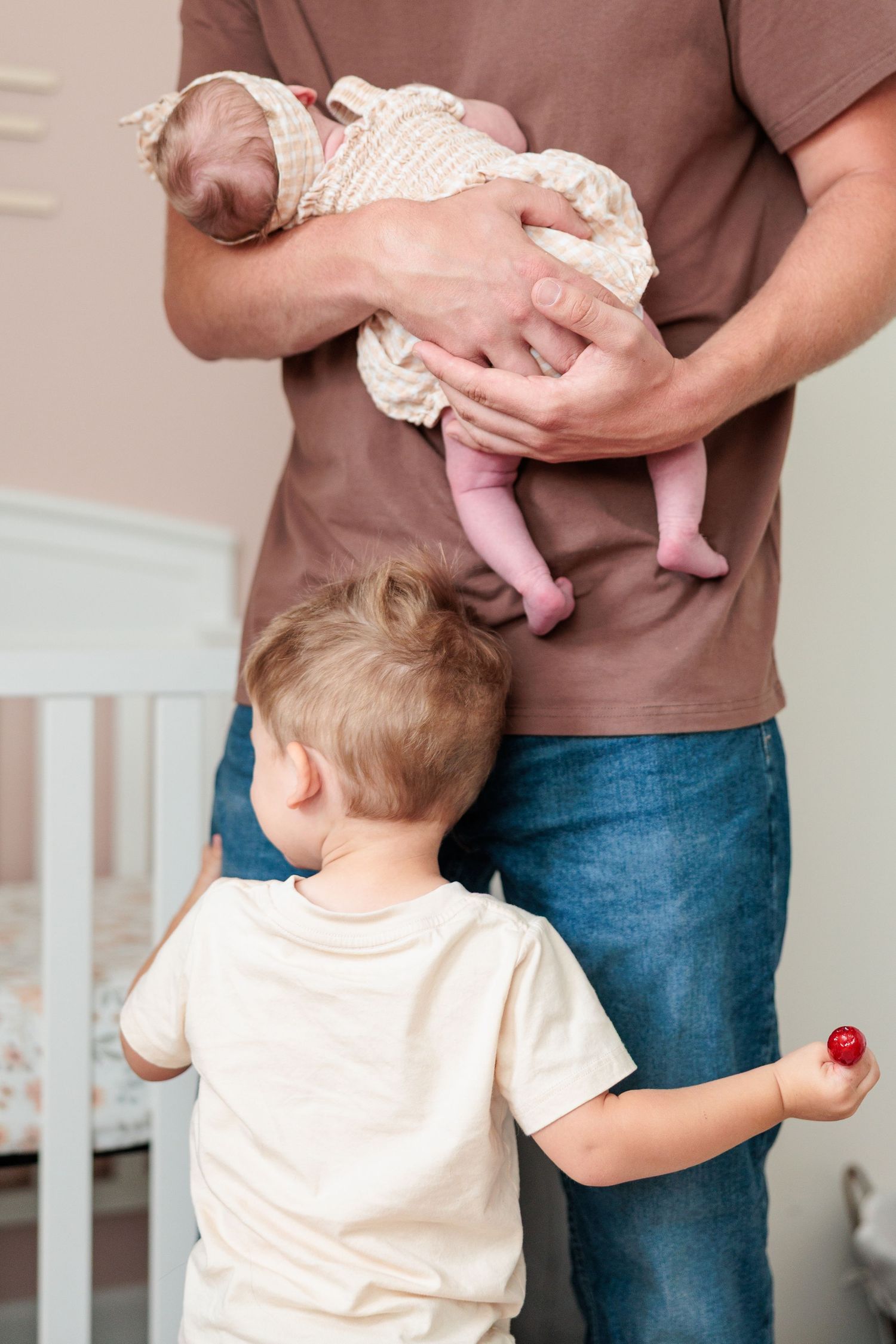 Father in brown shirt holds newborn baby while young toddler son stands beside him in nursery.