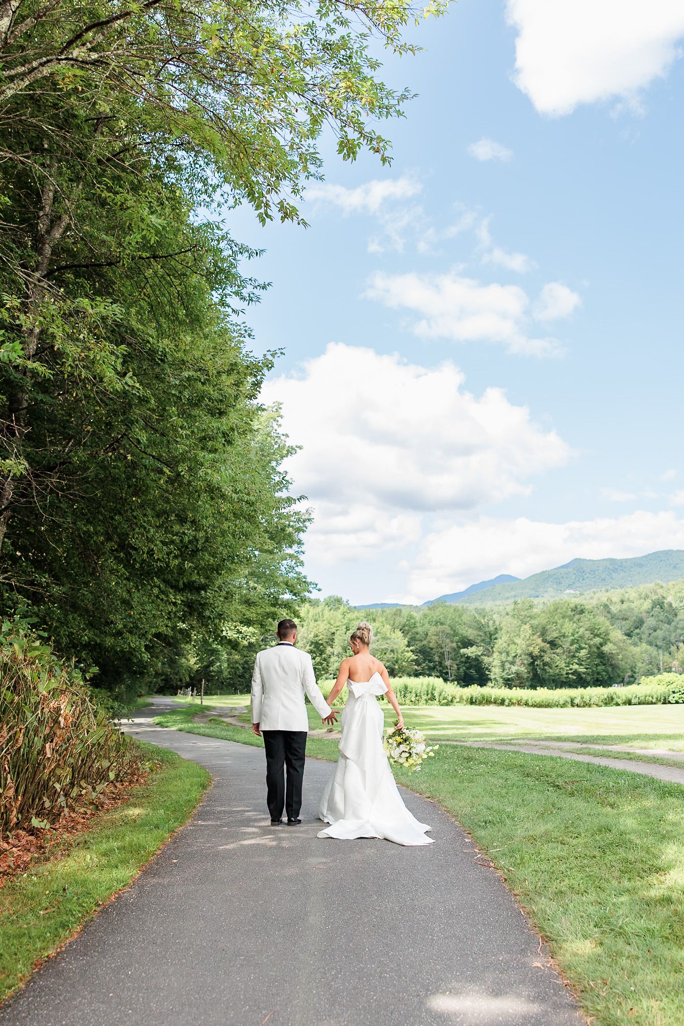 Bride and groom walk hand-in-hand down a scenic path with mountain views and lush green landscape.