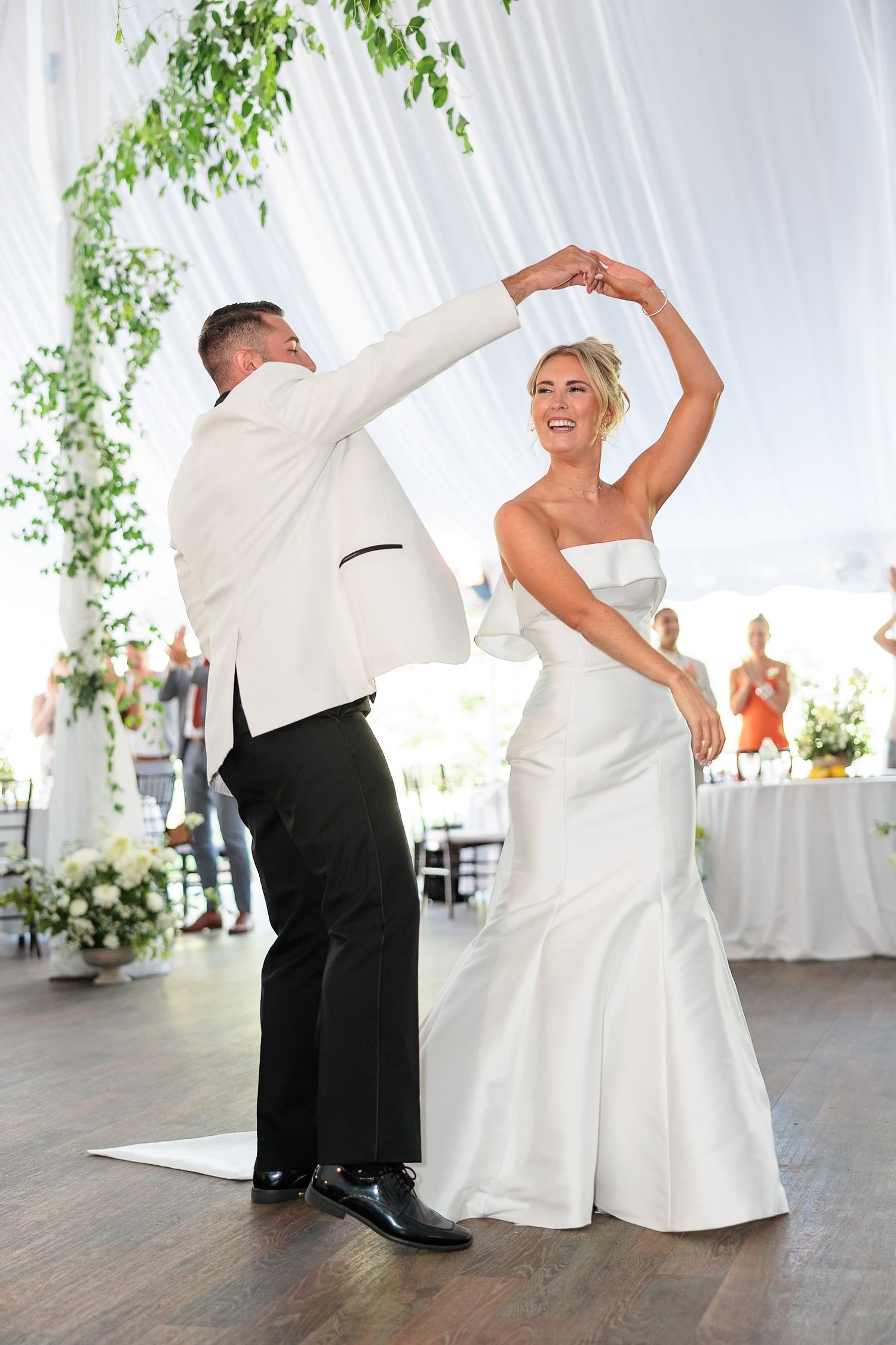 Bride and groom share first dance in white tuxedo and strapless gown under draped tent with greenery decor.