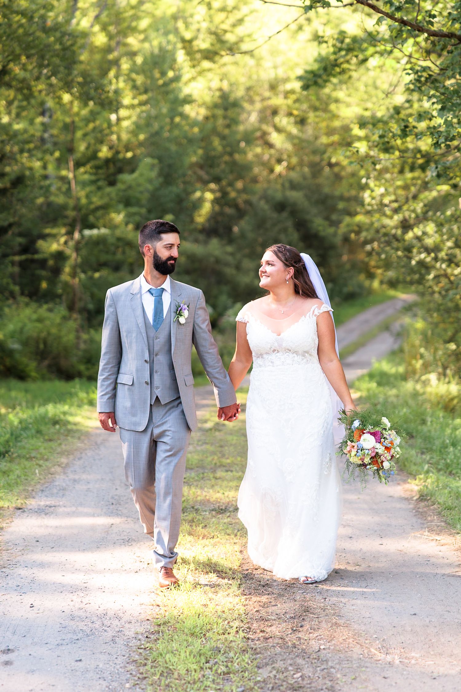 Bride and groom walk hand-in-hand down a sunlit tree-lined path, bride carrying a colorful bouquet.