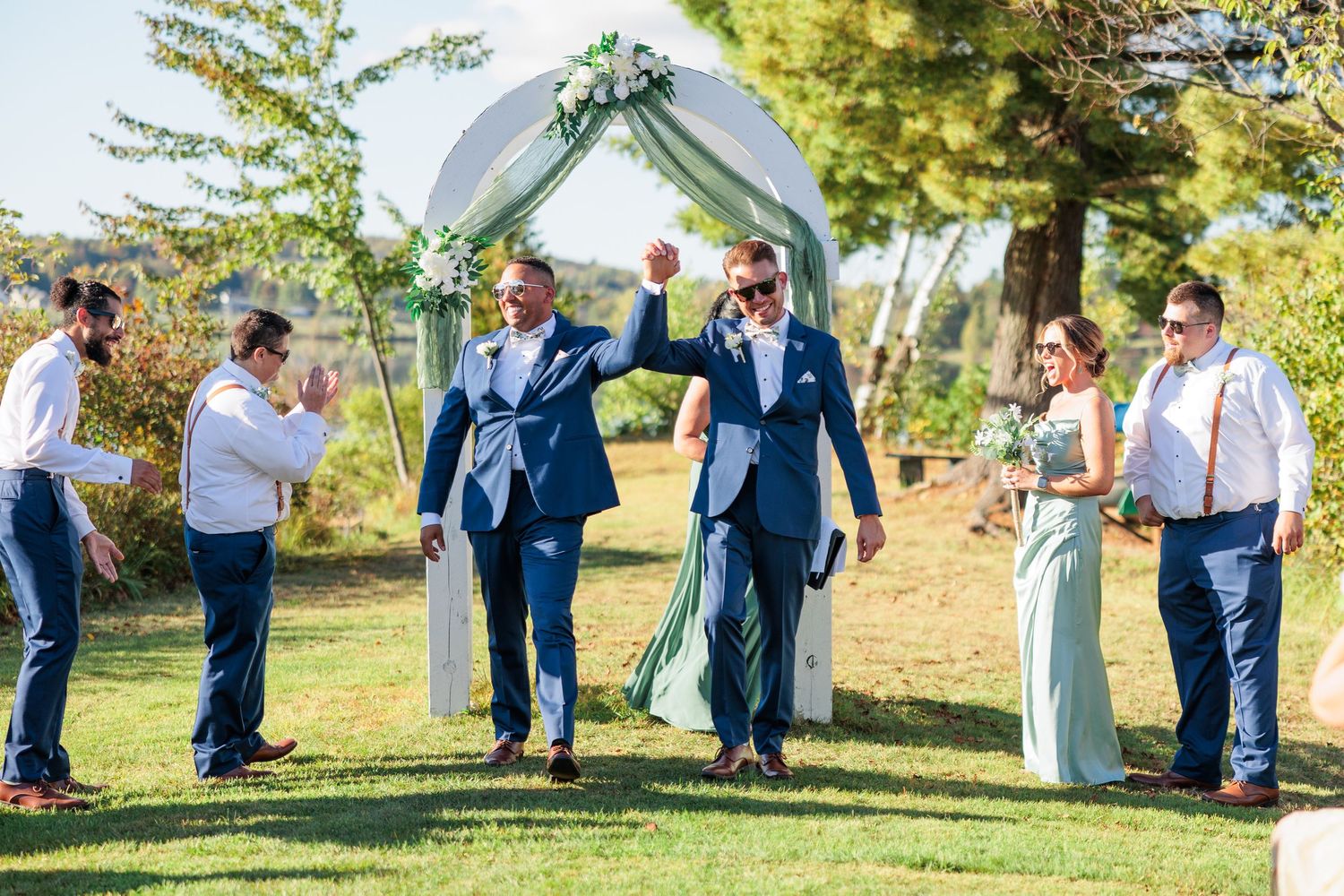 Wedding guests walk through an outdoor ceremony site with a white arch and chairs set up on green grass.