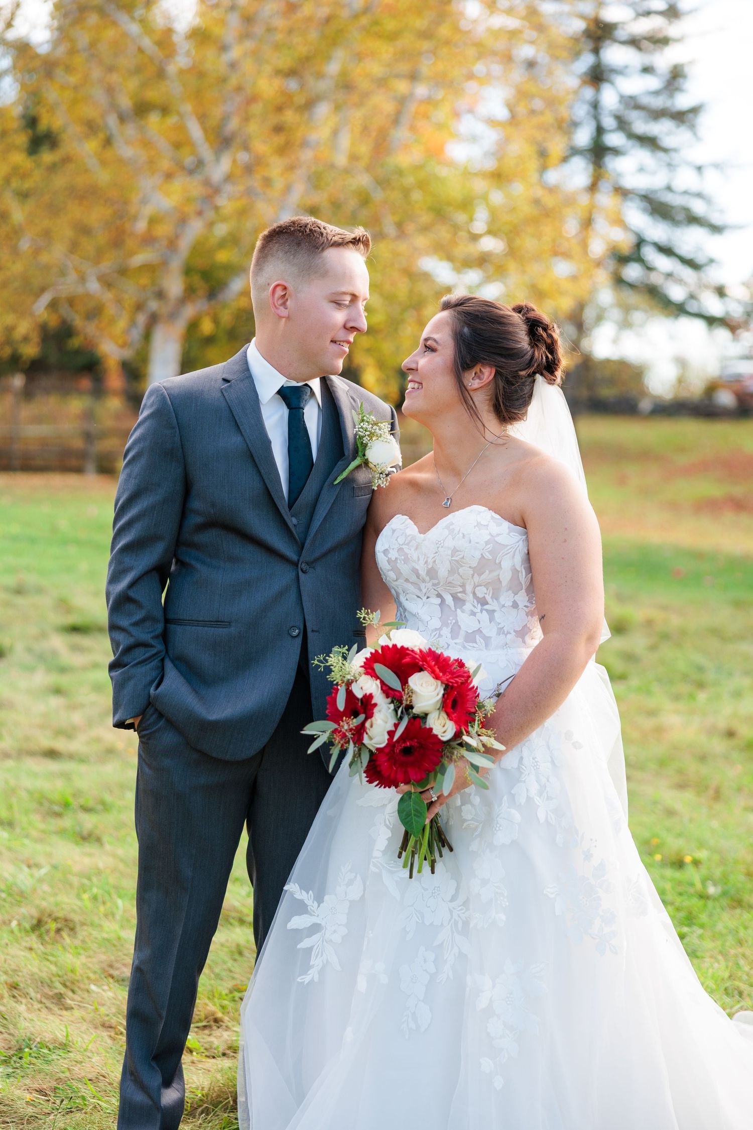 A romantic fall wedding portrait with a stunning red bouquet and elegant attire against golden autumn foliage.