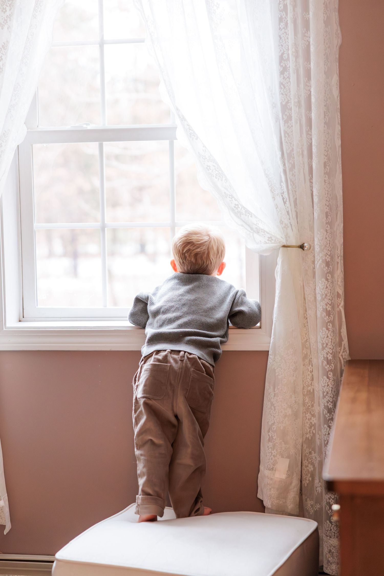 A child in grey clothing stands precariously on a window sill next to white curtains.
