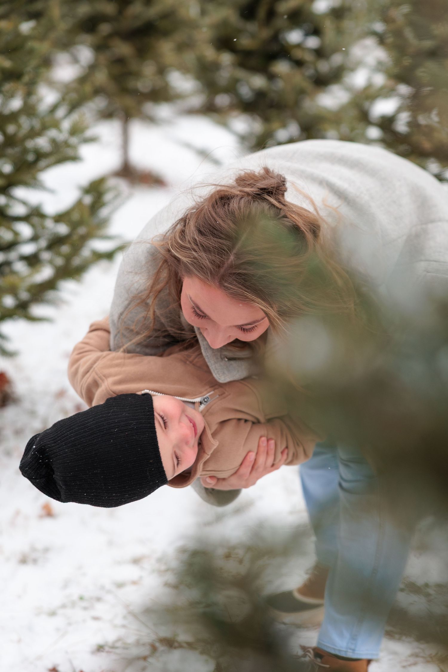 A playful couple embraces and laughs together among snow-covered Christmas trees outdoors.