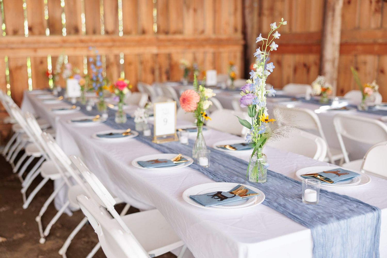Rustic barn wedding reception tables set with blue runners, wildflower centerpieces, and white folding chairs.