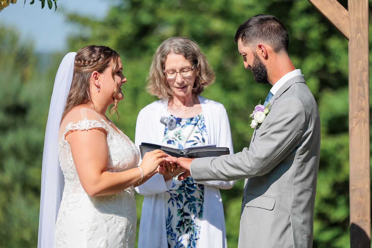 Bride and groom exchange vows with smiling officiant at outdoor wedding ceremony under blue sky.