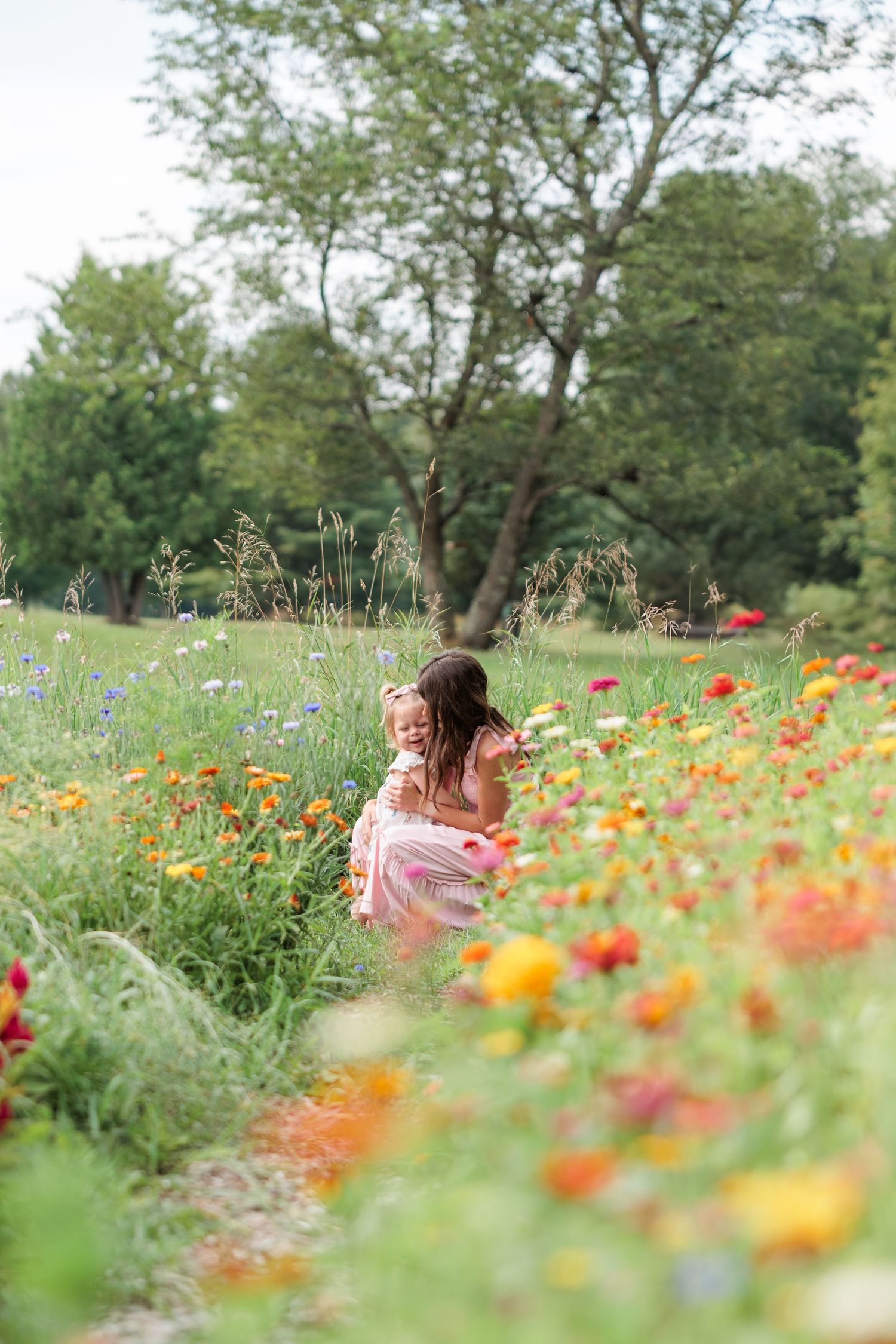 A mother and young daughter walk through a lush field of colorful wildflowers on a summer day.